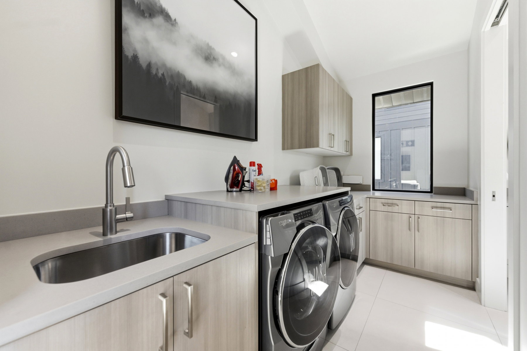 Dedicated laundry room featuring light wood-finish cabinetry, light gray countertops, a stainless steel undermount sink, and a black-framed window