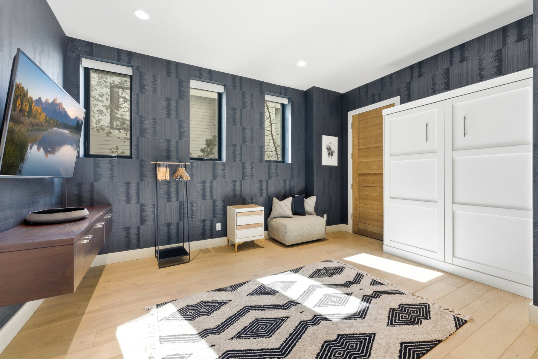 Room featuring light wood-finish flooring, two rectangular windows, a wall-mounted media console, and a built-in white cabinetry unit