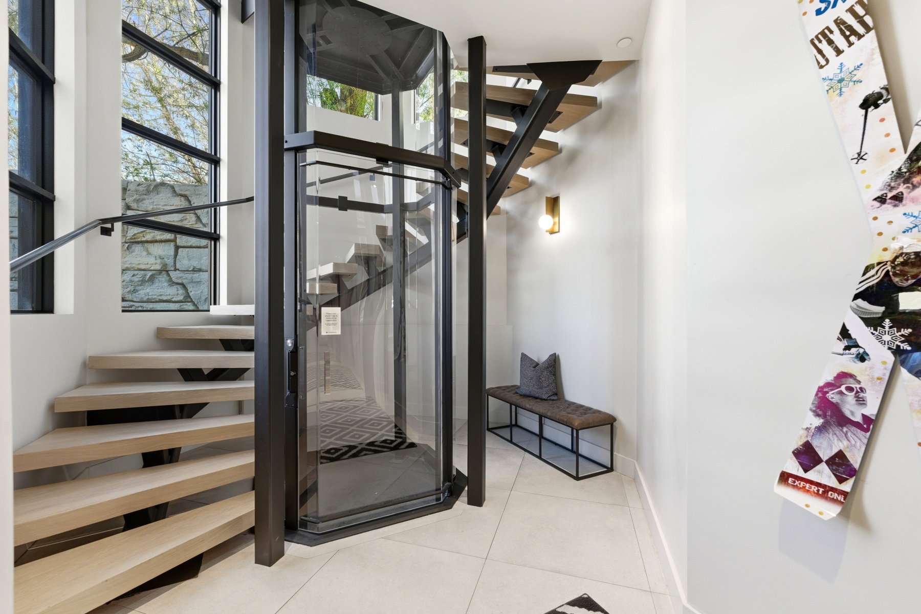 Contemporary interior featuring a glass-enclosed elevator, an open-tread staircase with light wood treads and dark railings, and large multi-pane windows