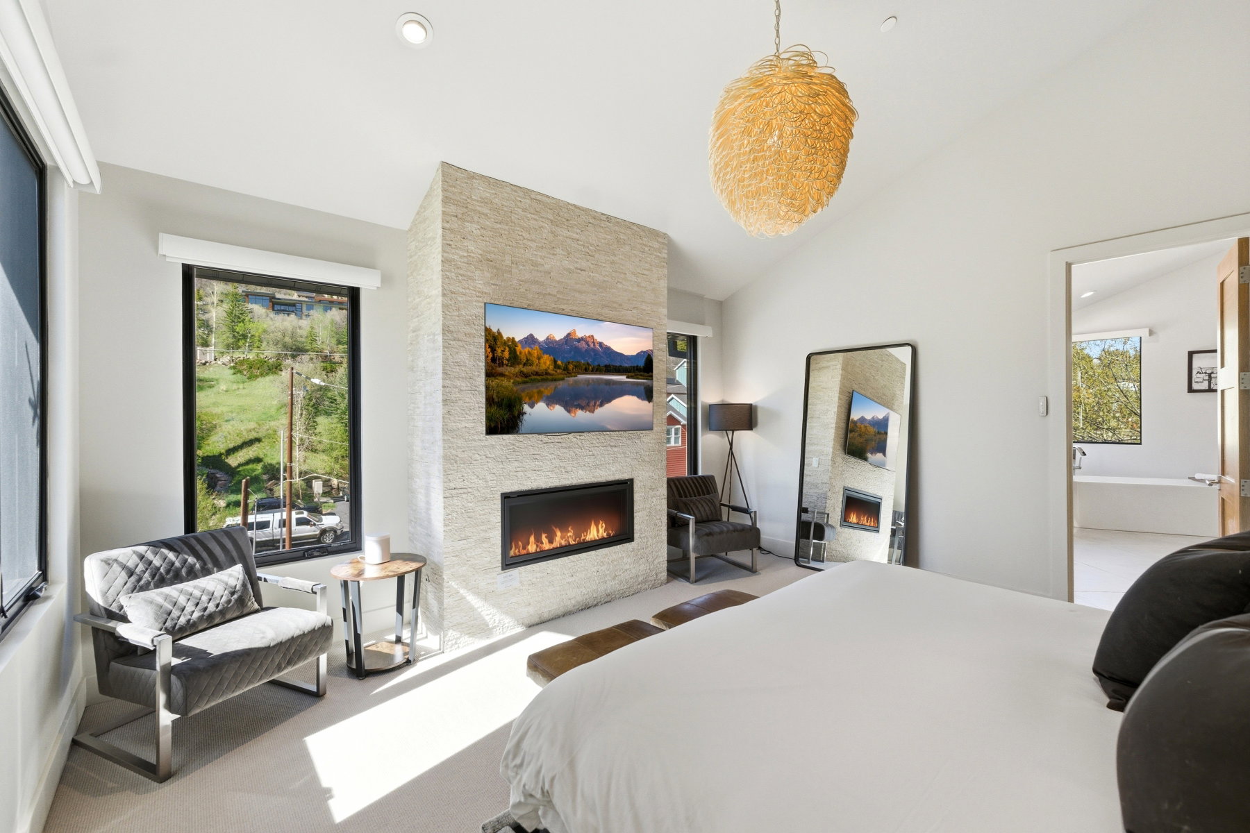 Vaulted ceiling bedroom featuring a stone-clad fireplace wall with an integrated linear fireplace, a wall-mounted television, and a large window with roller shades