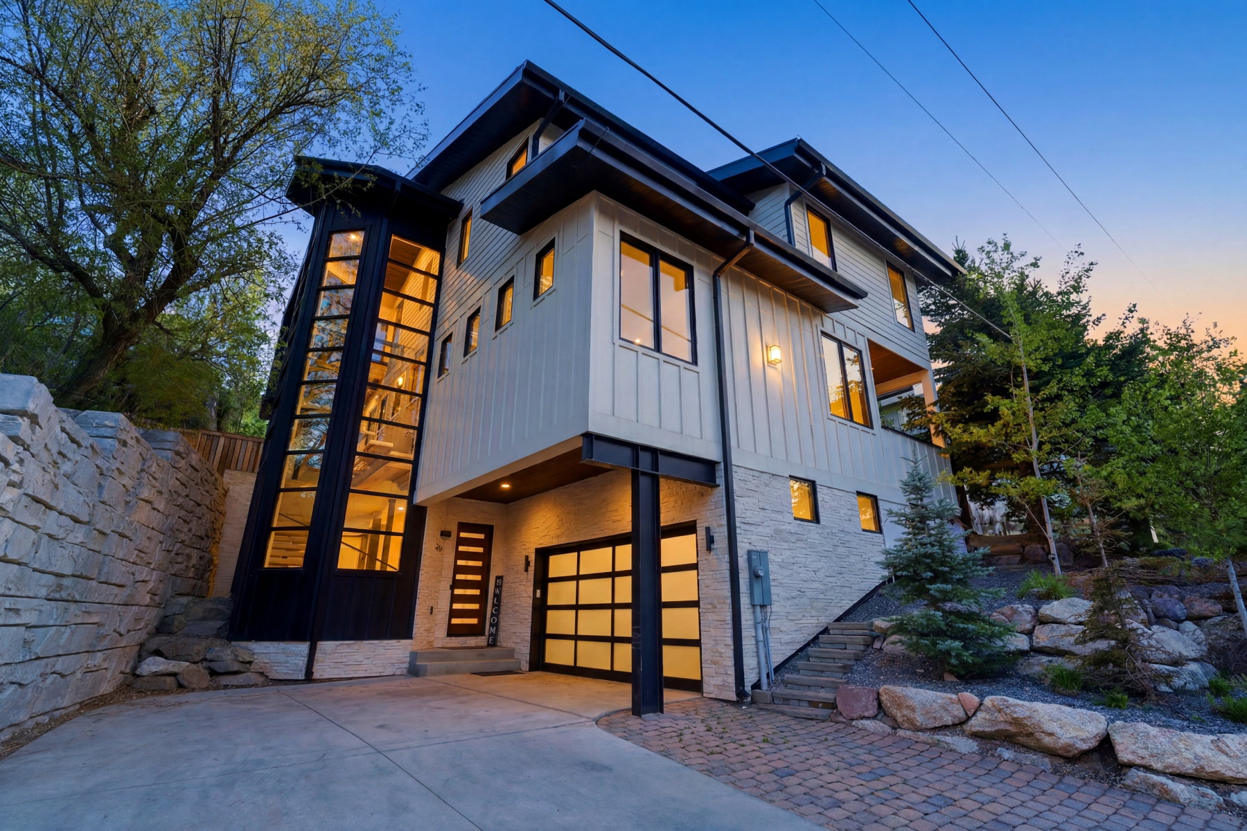 Modern architectural residence featuring a prominent glass-enclosed stairwell, light-colored vertical siding, and a stone retaining wall