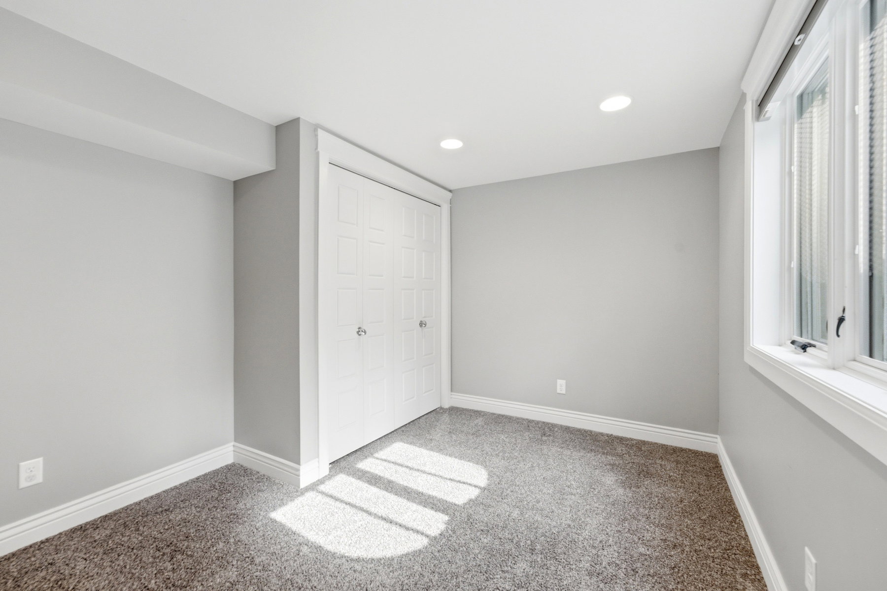 Soft gray wall finish in a room featuring a textured carpet, white bi-fold closet doors, recessed ceiling lights, and a white-framed window