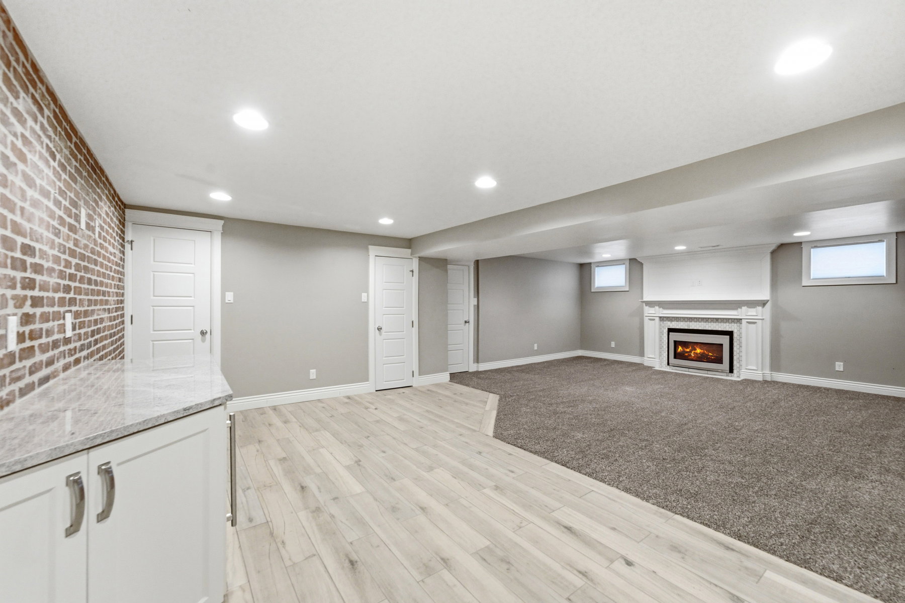 Spacious room featuring a white wood-finish built-in wet bar with a light stone-finish countertop, a brick accent wall, wood-finish flooring, recessed lighting, and a fireplace with a white mantel and surround