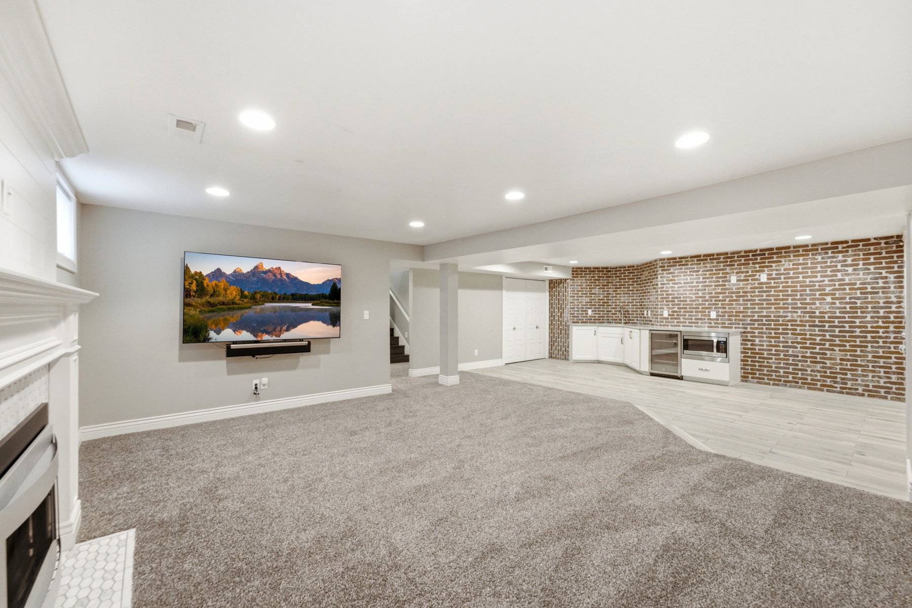 Spacious entertainment area featuring a white fireplace with hex tile surround, recessed lighting, and a built-in wet bar with white cabinetry, brick accent wall, and wood-finish flooring