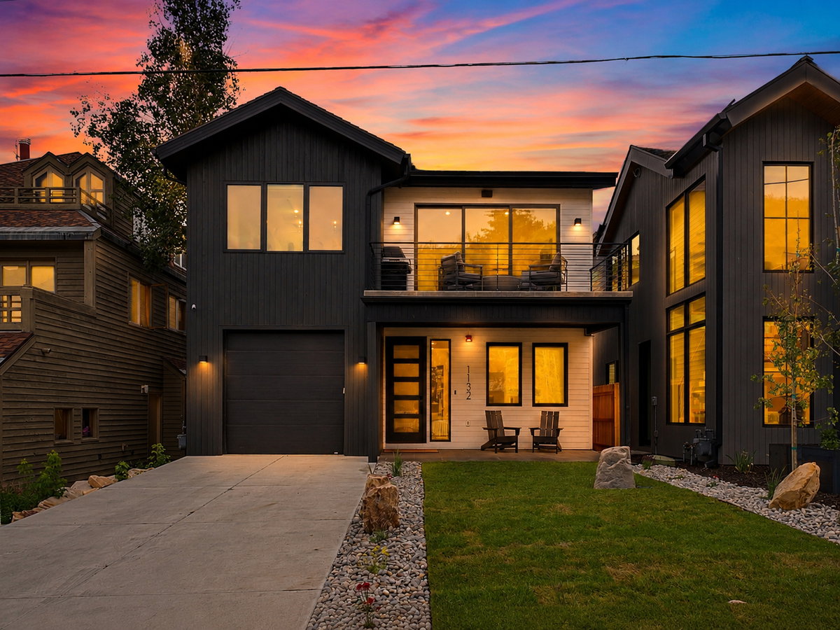 Modern two-story residence with a dark exterior, featuring a second-story balcony with metal railings and a spacious concrete driveway
