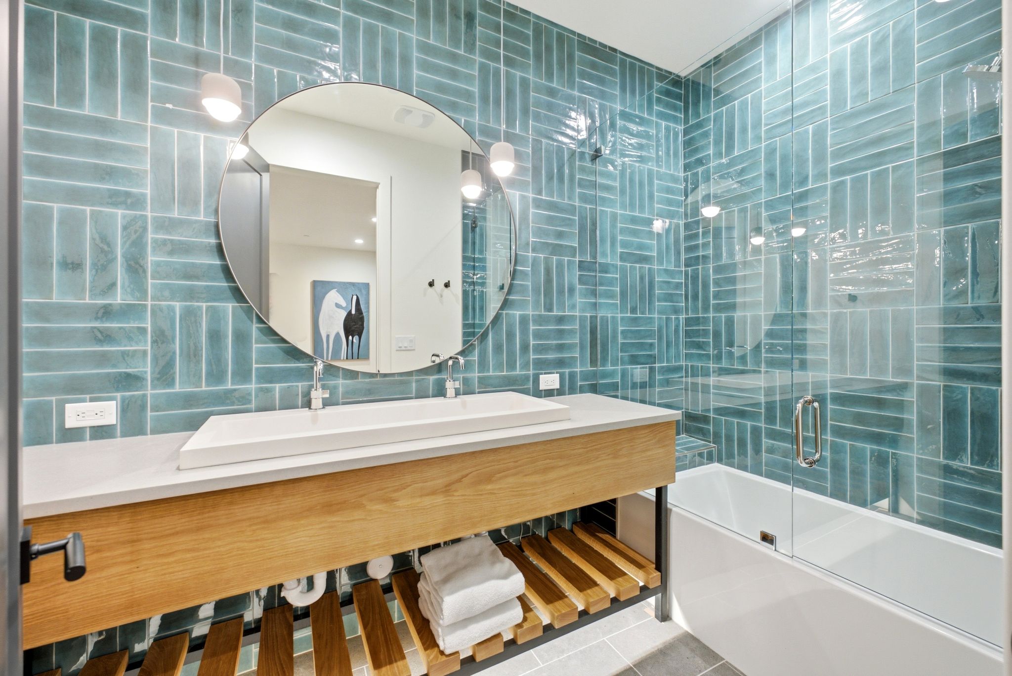 Bathroom featuring a deep soaking tub with a glass shower enclosure, blue subway tile, a rectangular vessel sink, a wood-finish vanity with open shelving, and a large round mirror
