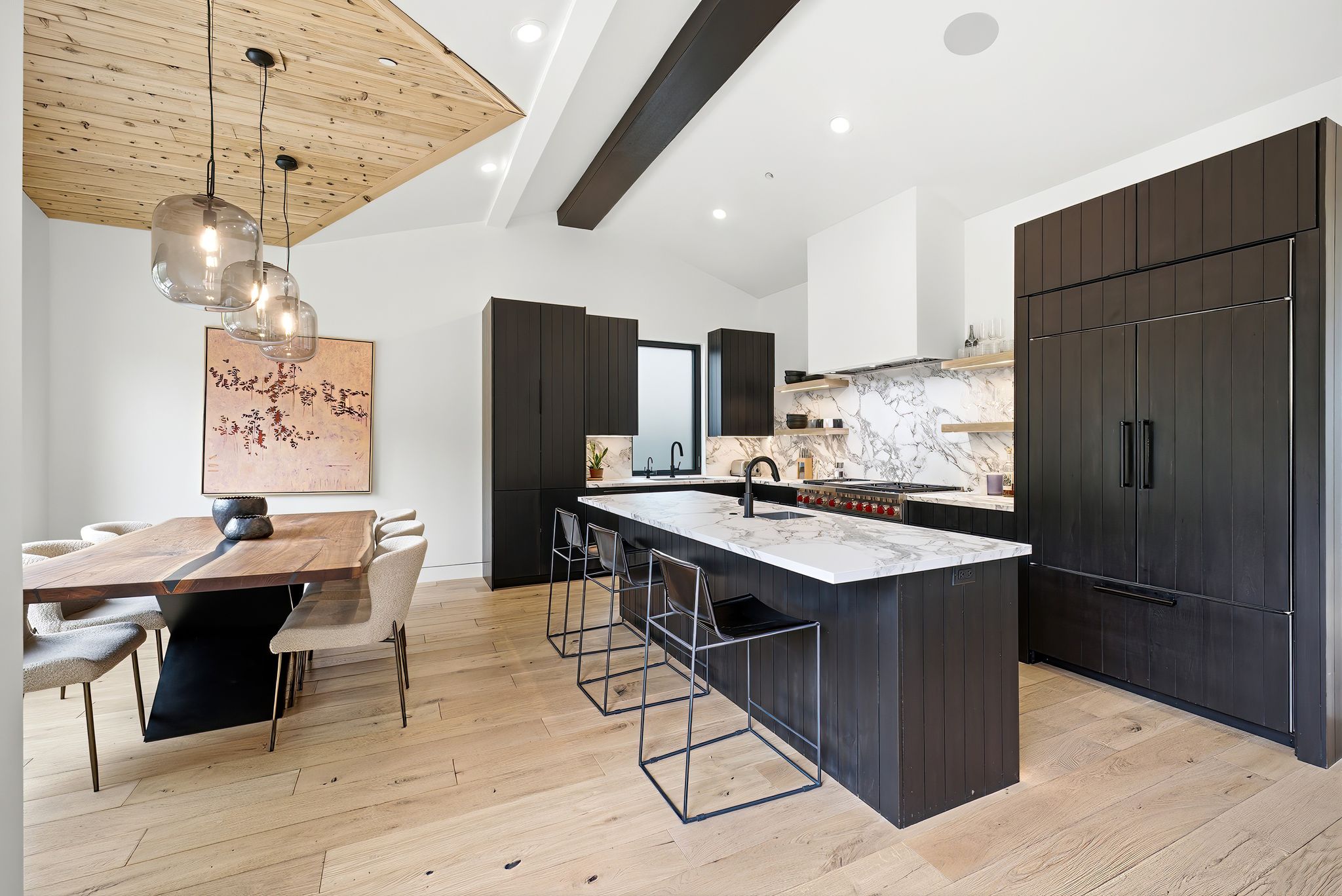 Contemporary kitchen and dining area featuring light wood-finish flooring, dark wood cabinetry, white vaulted ceilings, a large kitchen island with a light stone countertop, and a built-in paneled refrigerator