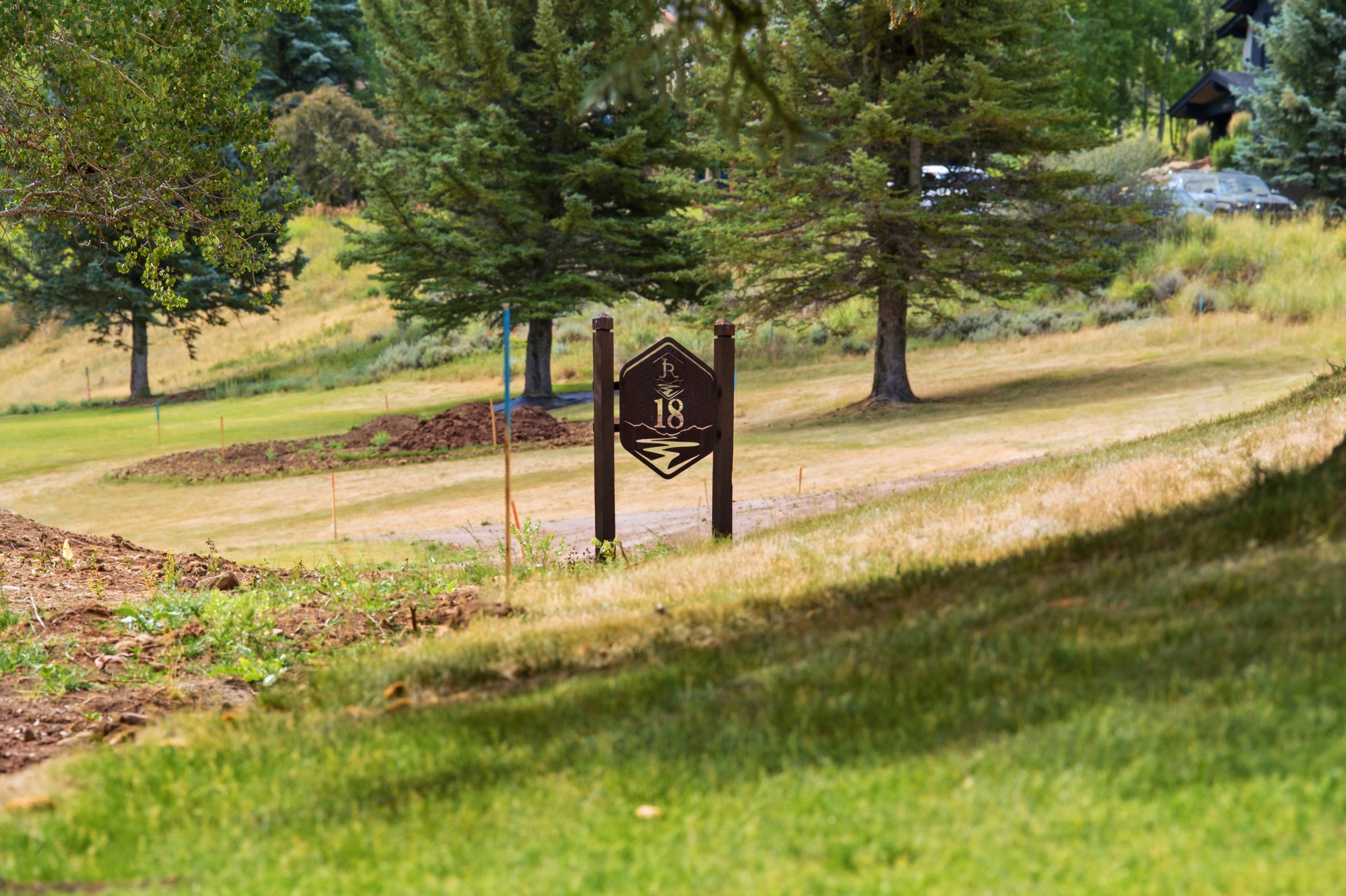 Golf course setting featuring a brown marker post, mature trees, a gently sloping grassy terrain, and distant residential structures