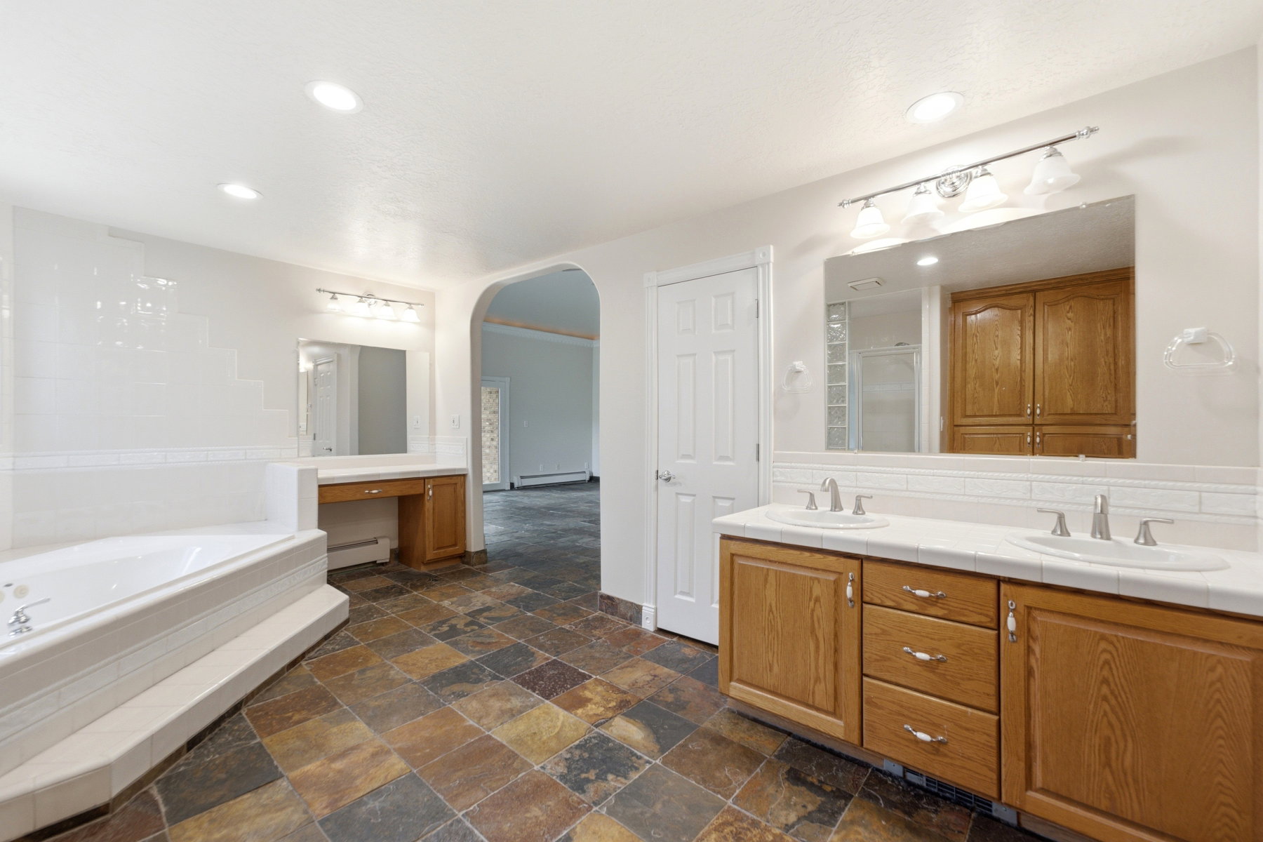 Spacious bathroom featuring a dual vanity with oak cabinetry and white countertops, a large jetted tub with tile surround, and an arched doorway leading to an additional space