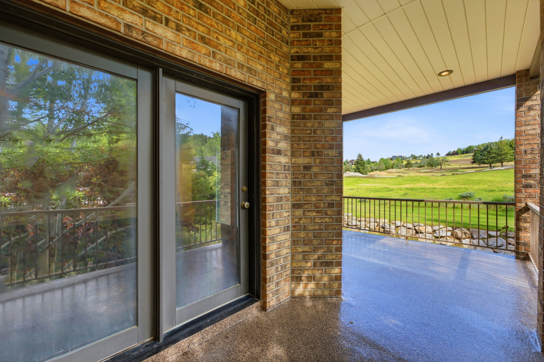 Covered outdoor patio featuring a textured floor, brick walls, and a metal railing overlooking a grassy landscape