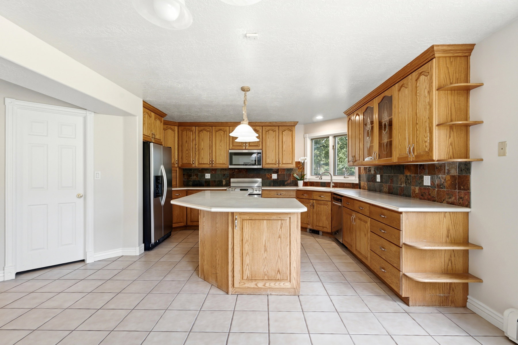 Kitchen featuring wood cabinetry, an angled island with a light-toned countertop, a tile backsplash, stainless steel appliances, and tile flooring