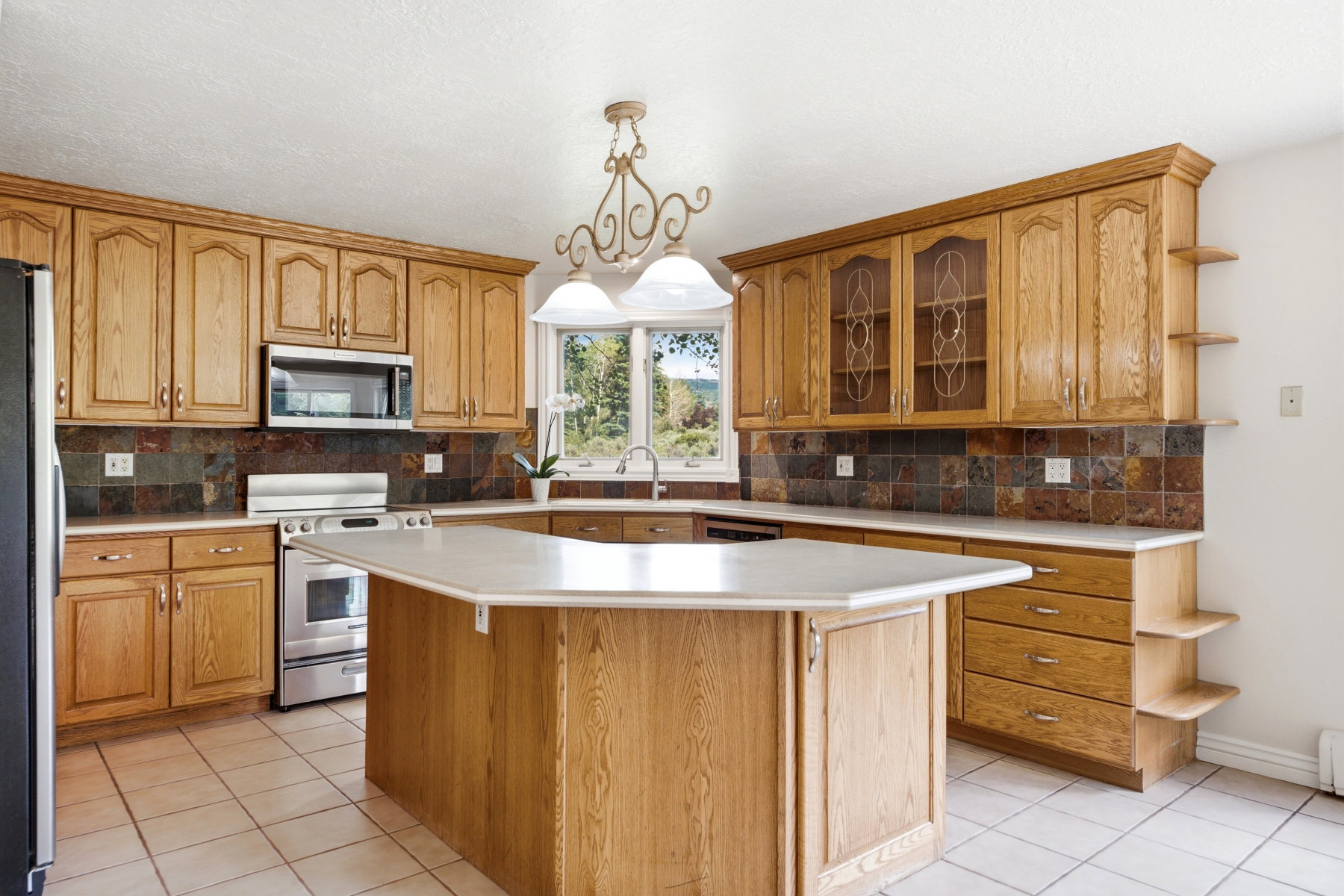 Kitchen featuring a central island with countertop space, light wood-finish cabinetry, stainless steel appliances, tile flooring, and a multi-toned tile backsplash