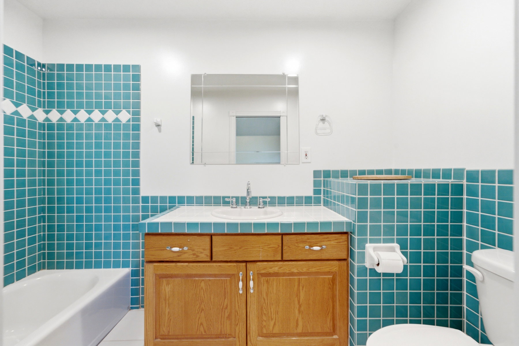 Bathroom featuring teal square wall tiling with a decorative white accent band, a white bathtub, a wood-finish vanity with a built-in sink, and a framed mirror