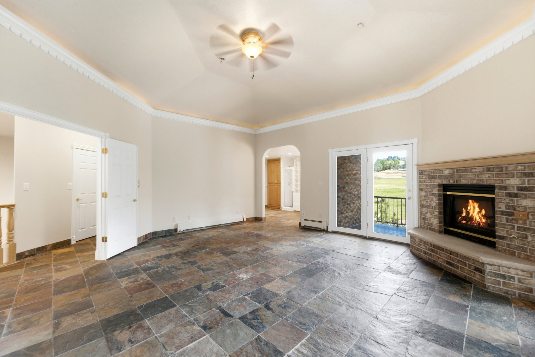 Spacious room featuring slate tile flooring, a brick fireplace with a hearth, double French doors, and a ceiling fan with integrated lighting