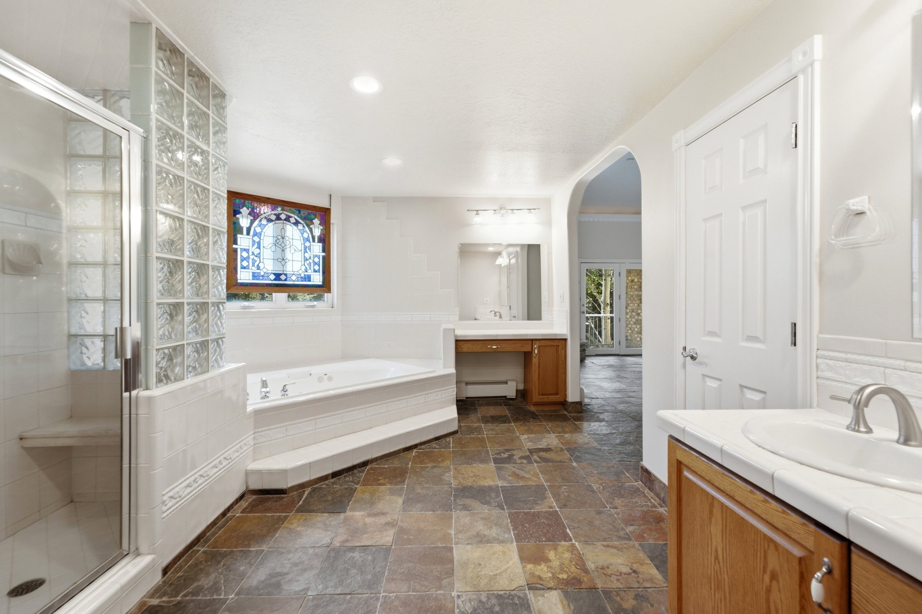 Spacious bathroom featuring natural stone flooring, a glass-enclosed shower with built-in bench, a jetted soaking tub, and dual vanity areas with solid surface countertops