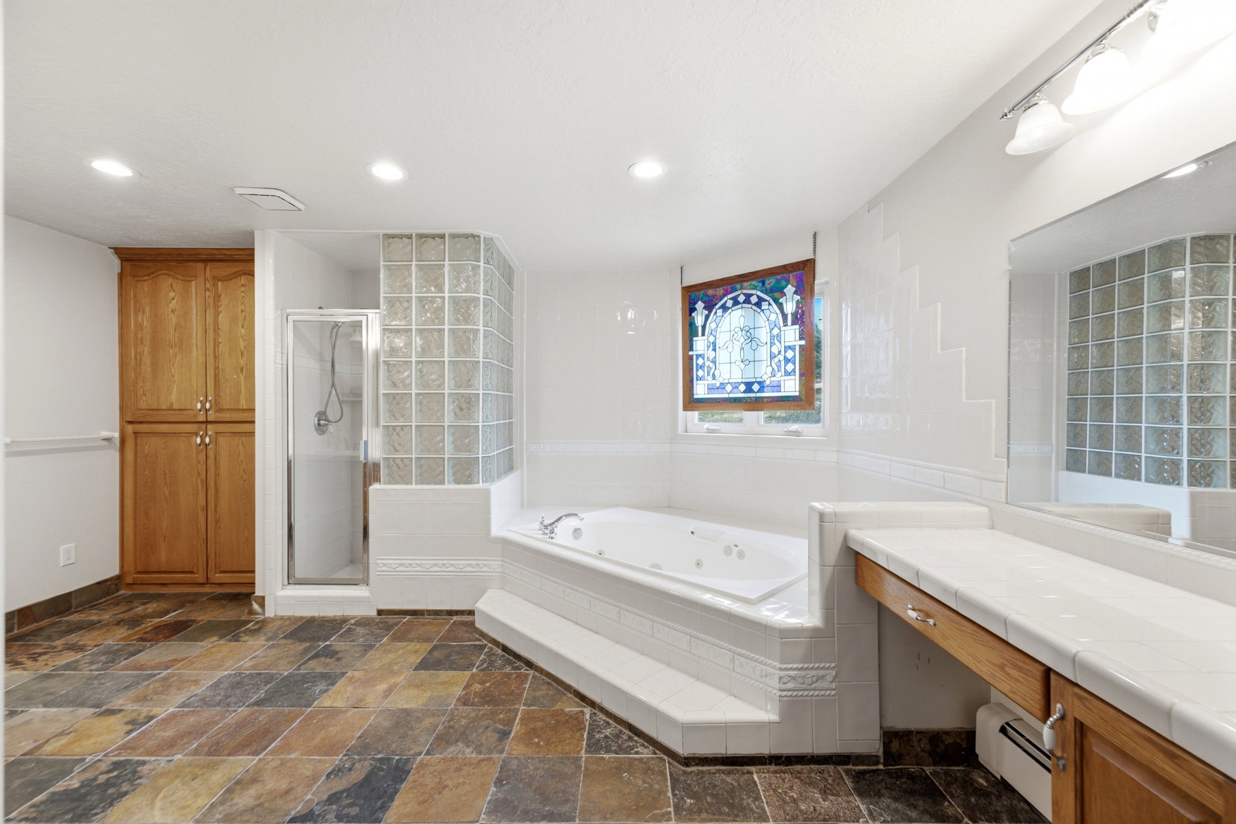 Spacious bathroom featuring a multi-toned slate tile floor, a built-in jetted tub, a glass-block shower enclosure, a stained-glass window, and a wood-finish vanity with a tile countertop