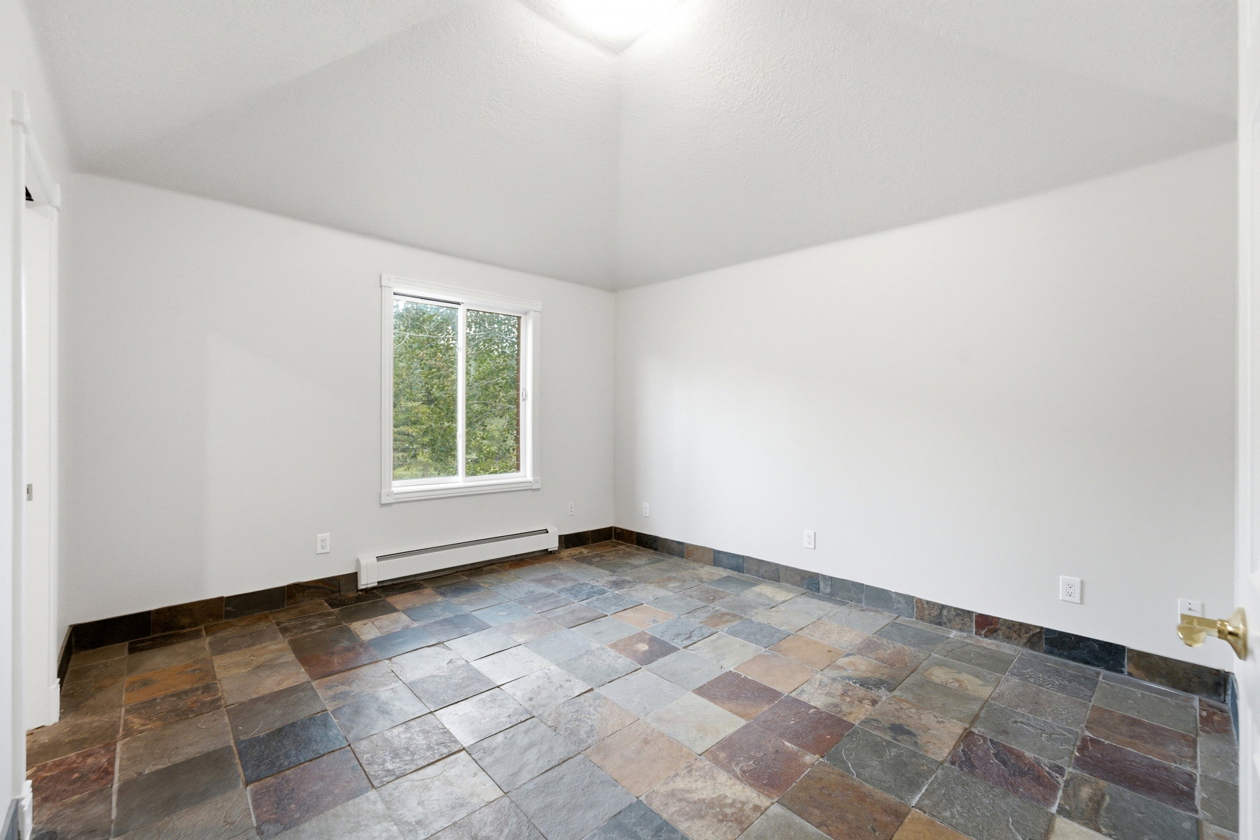 Spacious room featuring slate tile flooring, white walls, and a single window with a tree-lined exterior