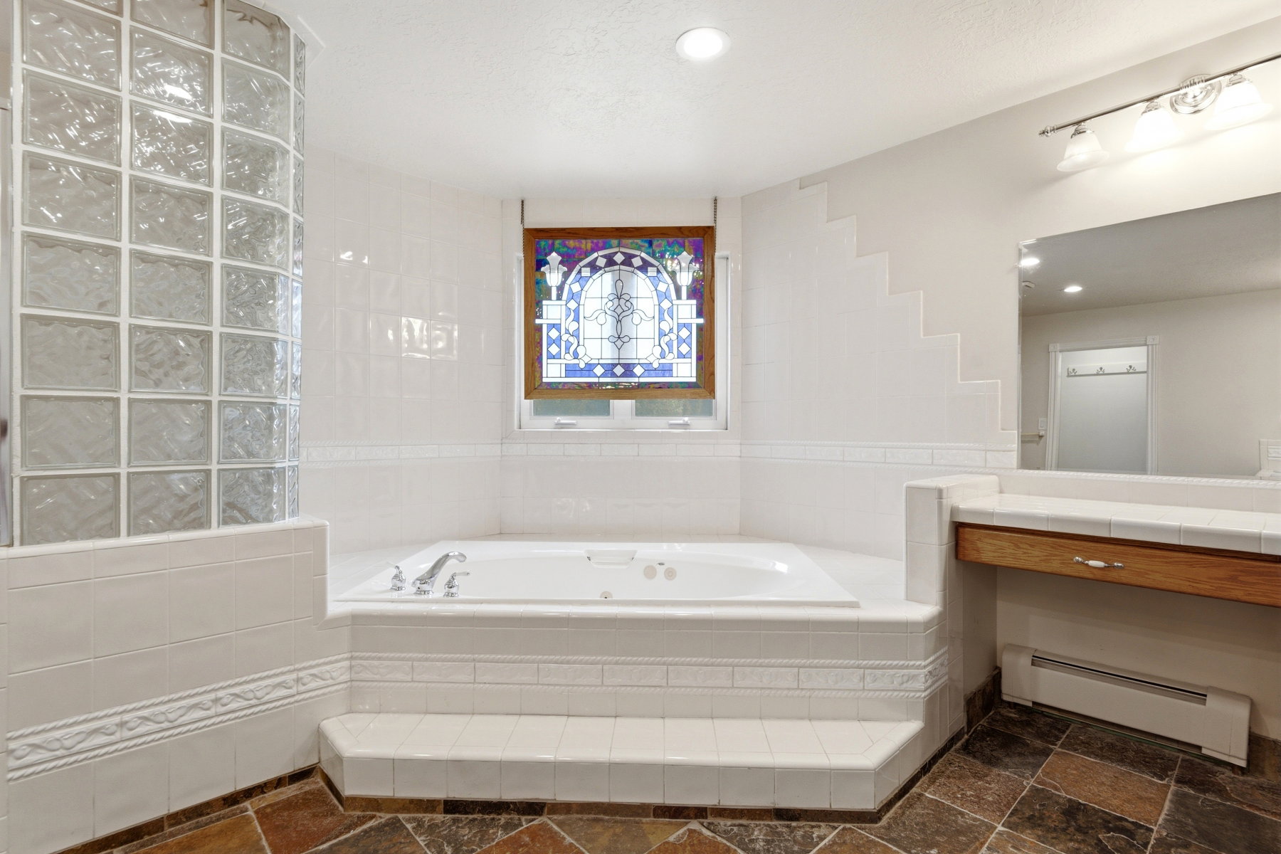 White tiled bath area featuring a built-in jetted tub, a stained-glass window, a glass block shower enclosure, a vanity with a wood-finish base, and slate-style floor tiling