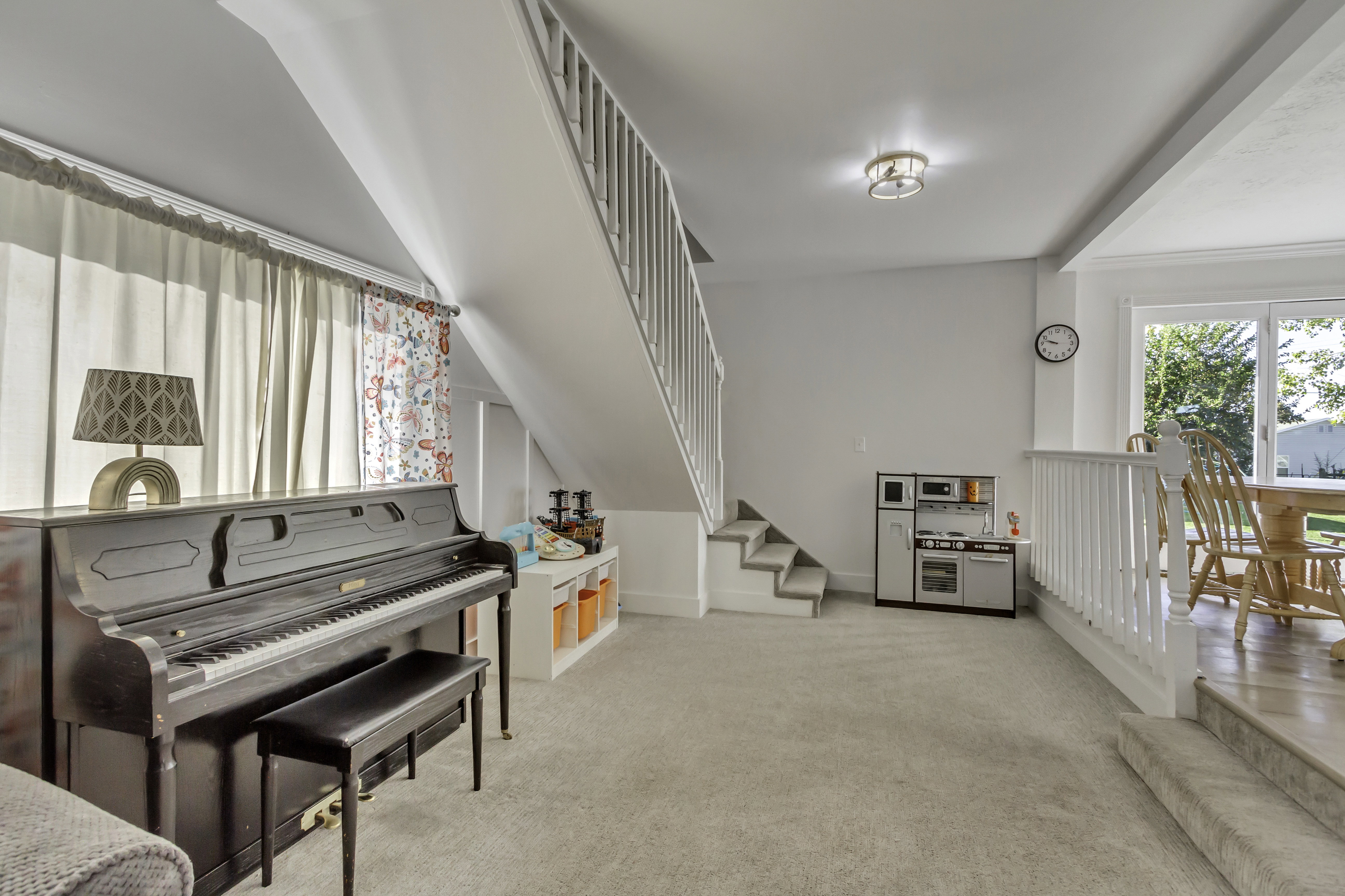 Carpeted lower-level room featuring a staircase with white balusters, recessed storage shelving, a window with exterior greenery, and a ceiling-mounted light fixture