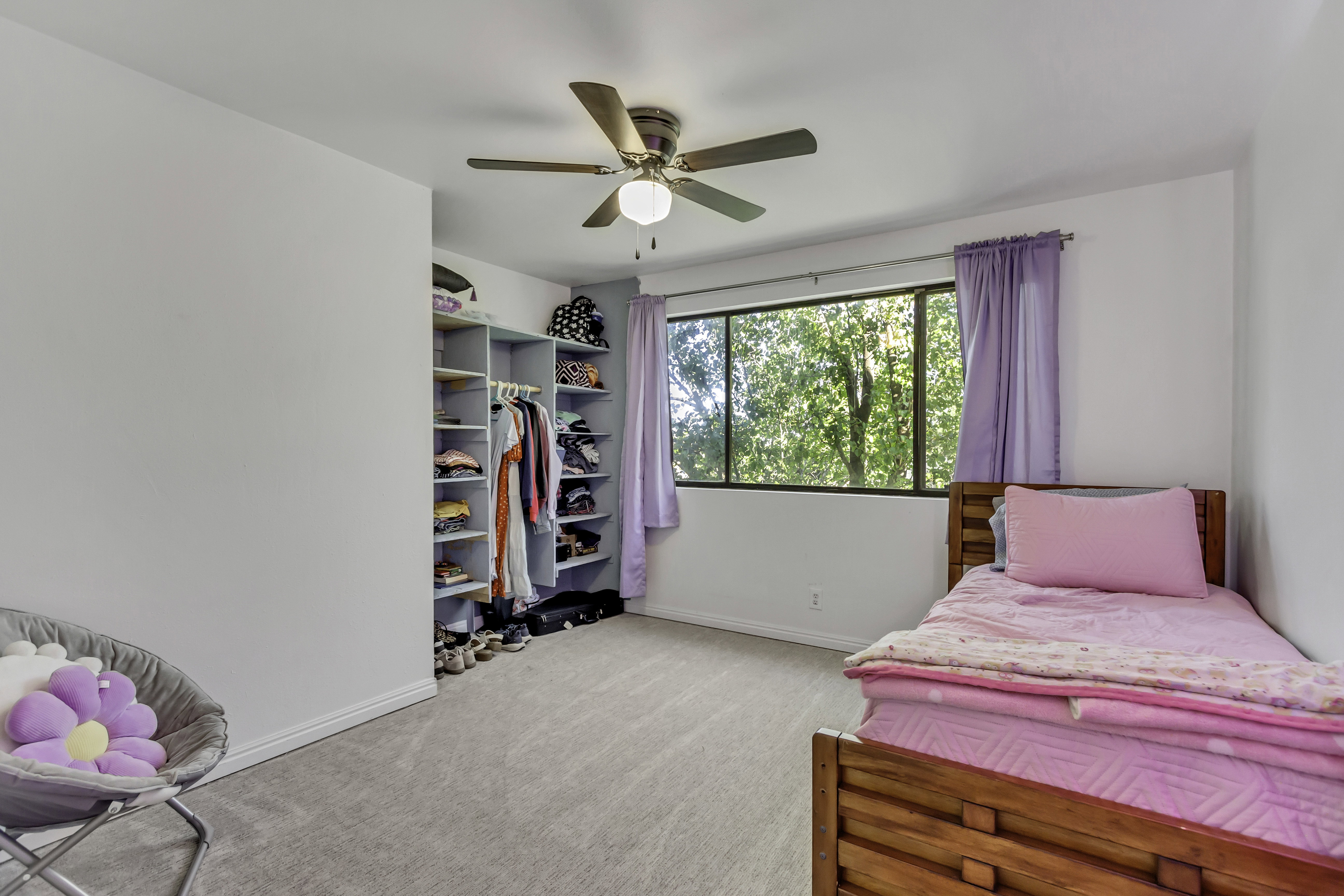 Carpeted room featuring a ceiling fan with integrated lighting, a large window with exterior foliage, and built-in closet shelving