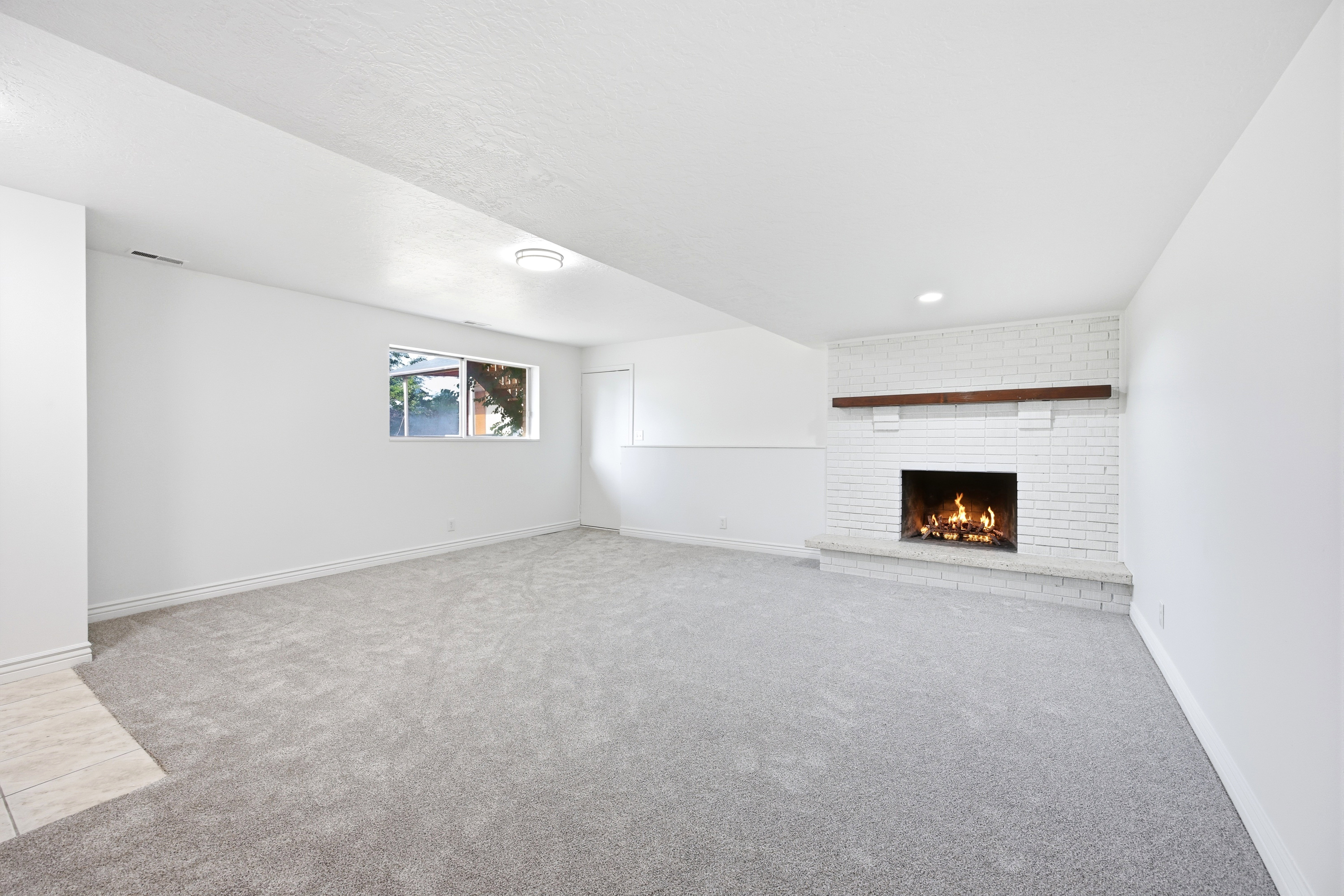 Spacious room featuring light gray carpeting, a white painted brick fireplace with a wood mantel, recessed lighting, and a window providing natural light