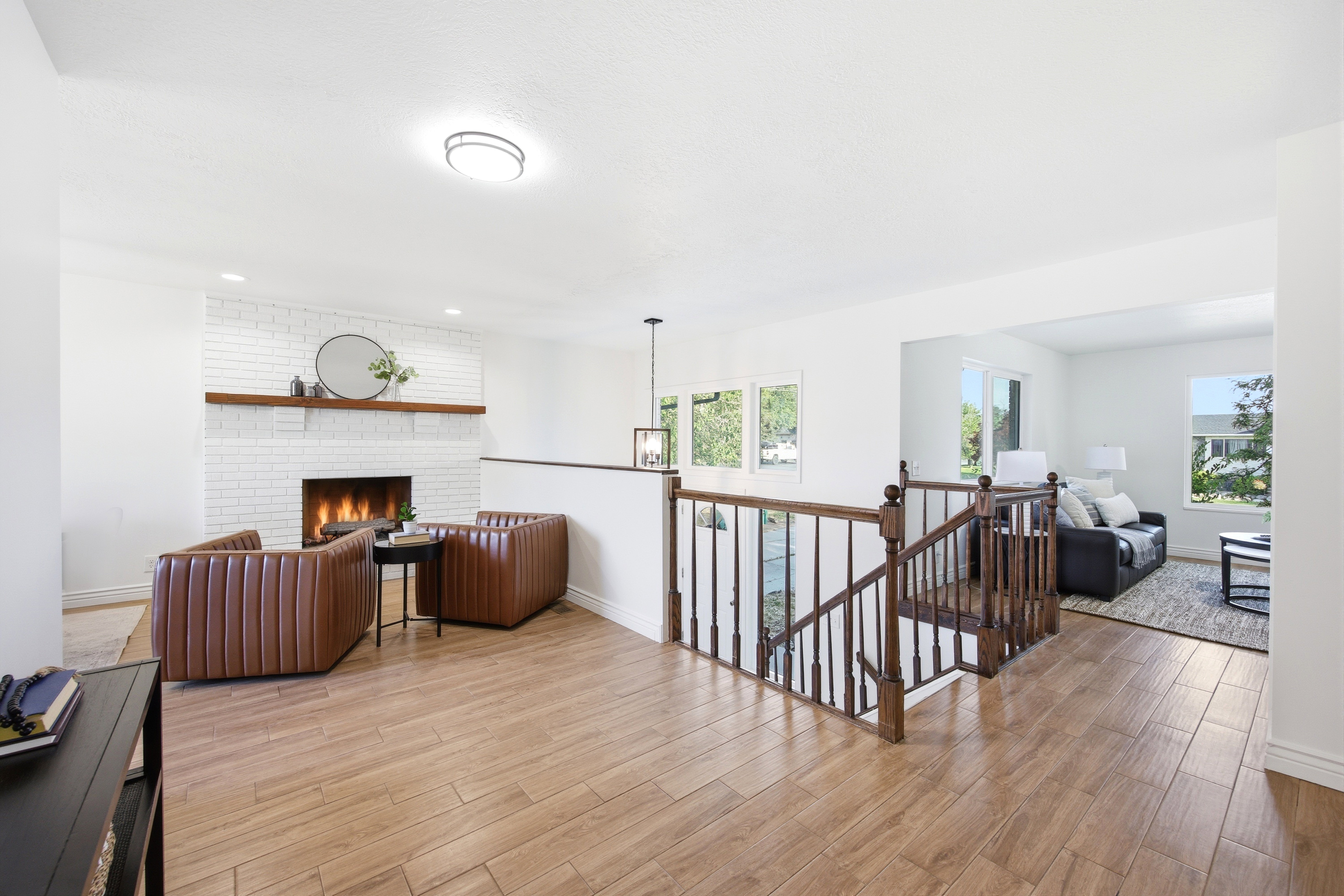 Expansive open-concept space featuring wood-finish flooring, a white brick fireplace with a wood mantle, and a dark wood staircase with wrought iron balusters