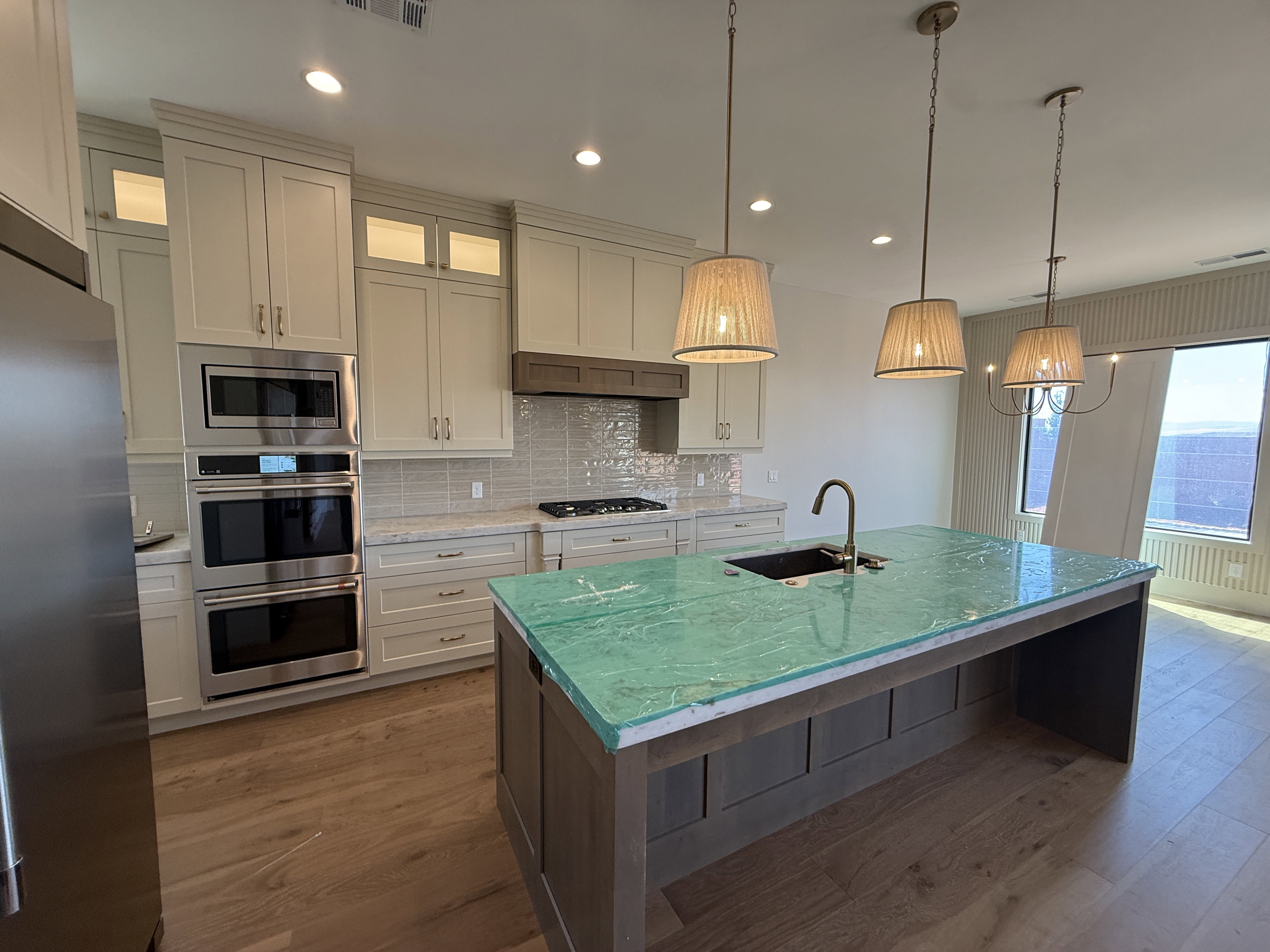 Spacious kitchen featuring an extended island with a distinctive green countertop, stainless steel appliances, light-toned cabinetry, wood-finish flooring, and recessed lighting