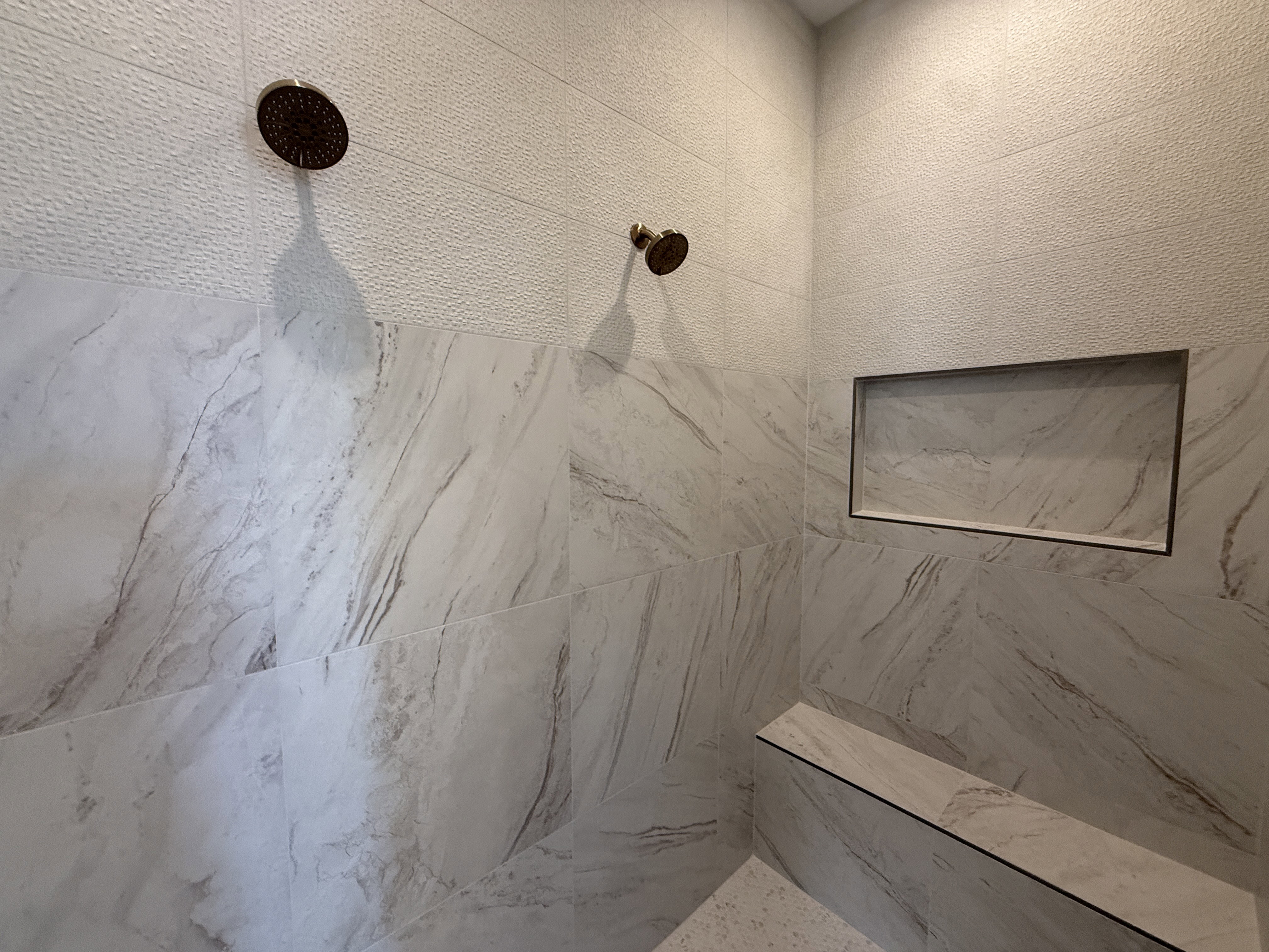 Spacious shower stall featuring dual bronze-finish showerheads, large format marble-look wall tiling, a built-in shower niche, and a stone-finish shower bench