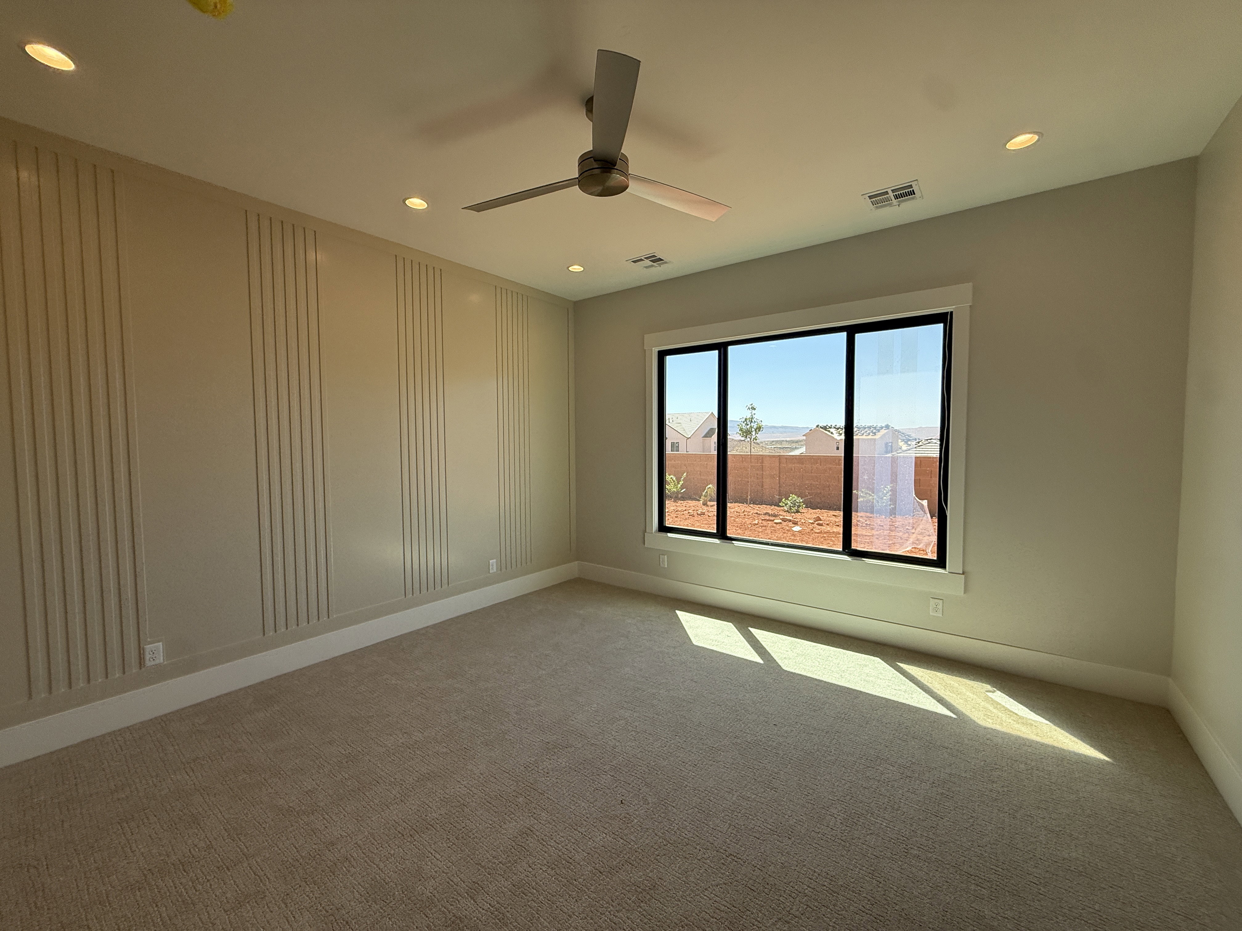 Spacious interior featuring a large window with black framing, textured accent wall paneling, ceiling fan, recessed lighting, and carpet flooring