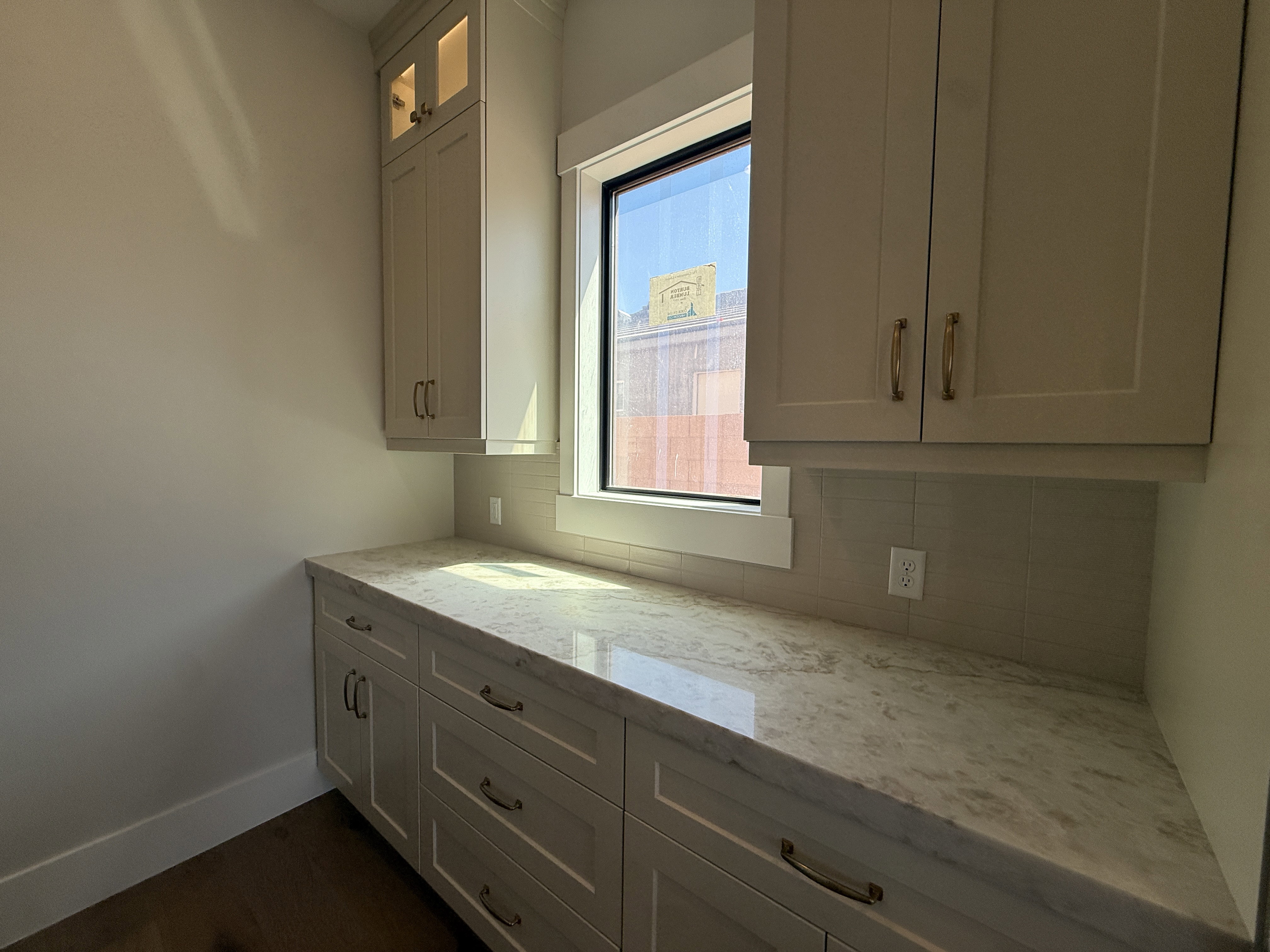 Light-filled space featuring extensive shaker-style cabinetry with gold-finish hardware, a substantial stone countertop, a window with dark trim, and a rectangular tile backsplash