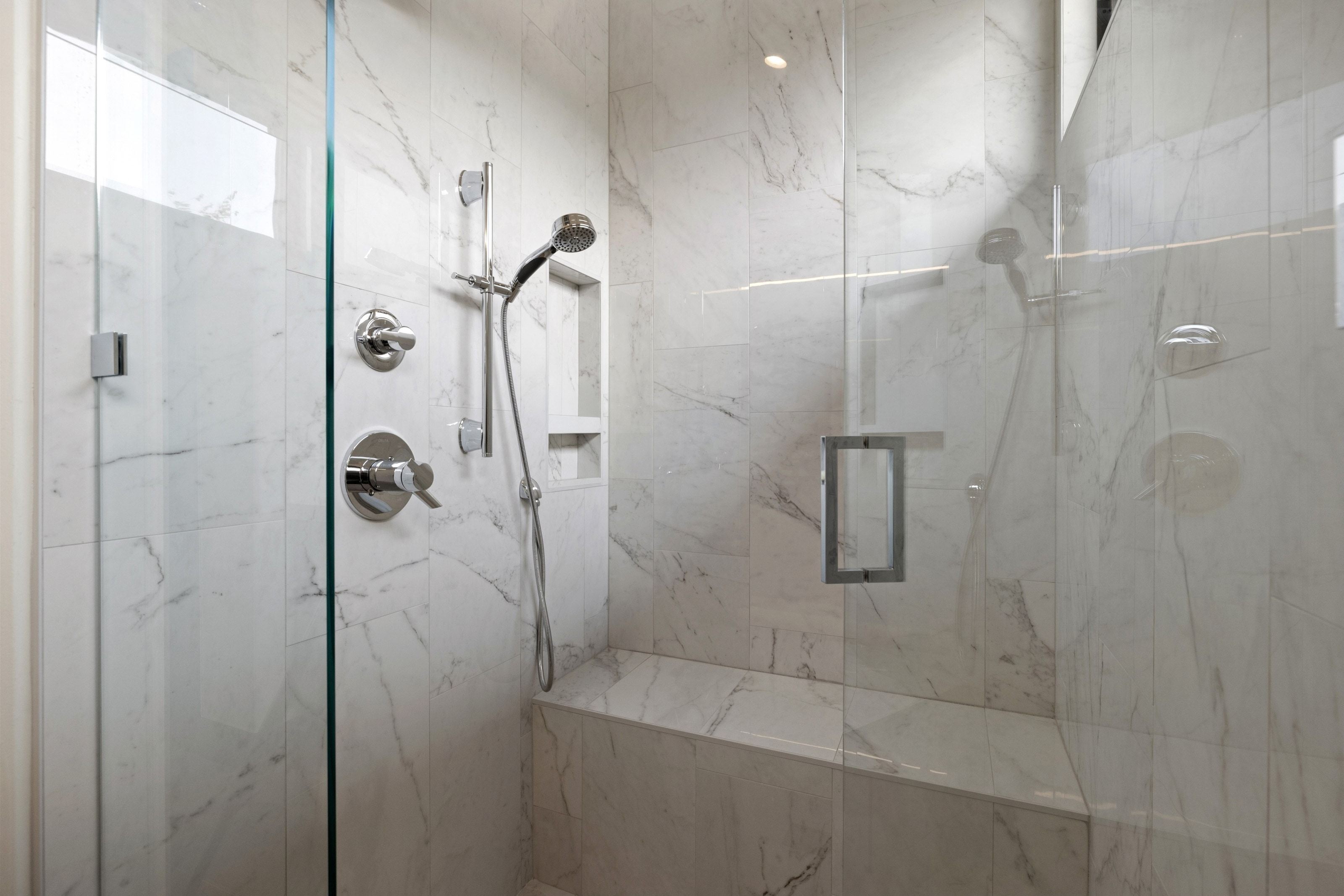 Walk-in shower featuring clear glass enclosure, marble-look tile walls, chrome fixtures, built-in bench, and recessed storage niche