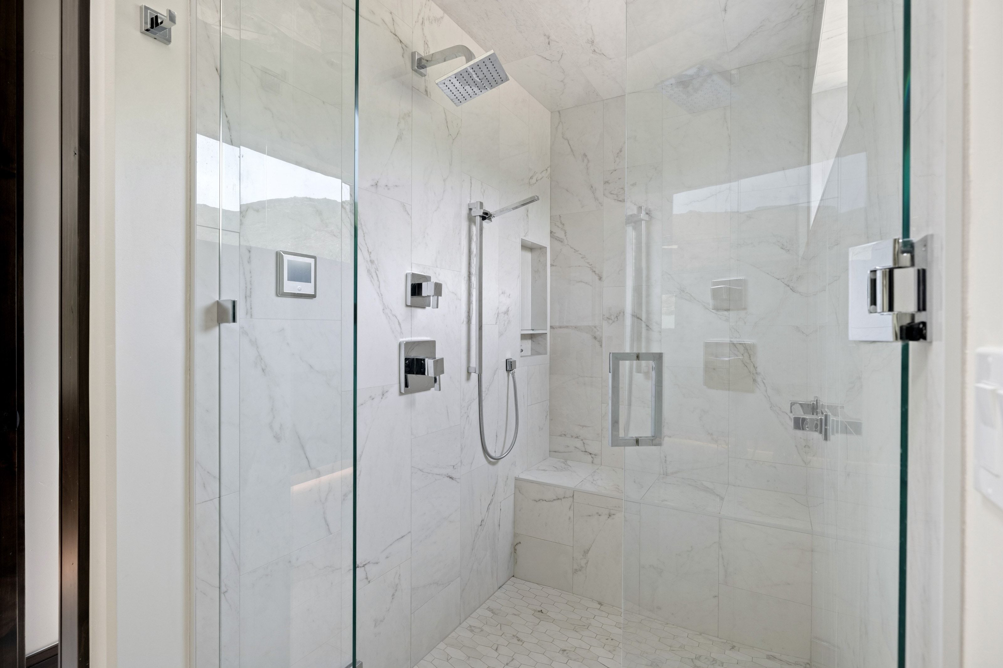 Glass-enclosed shower featuring large format white tiled walls with subtle veining, a built-in tiled bench, a rain showerhead, a handheld shower fixture, and a mosaic tiled floor