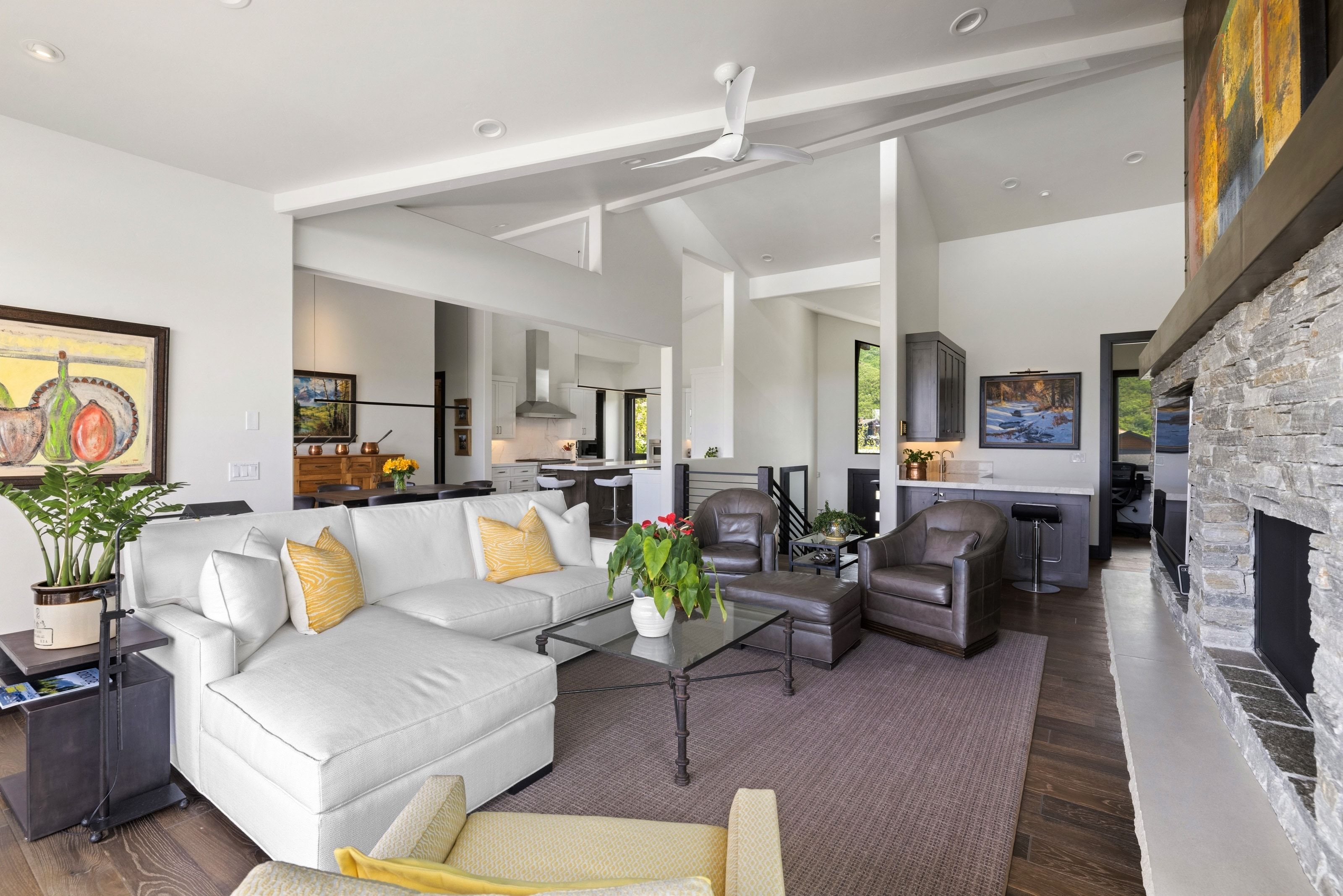 Open-concept living area featuring a stone-clad fireplace, vaulted ceilings with exposed beams, wood-finish flooring, a built-in wet bar with cabinetry, and recessed lighting