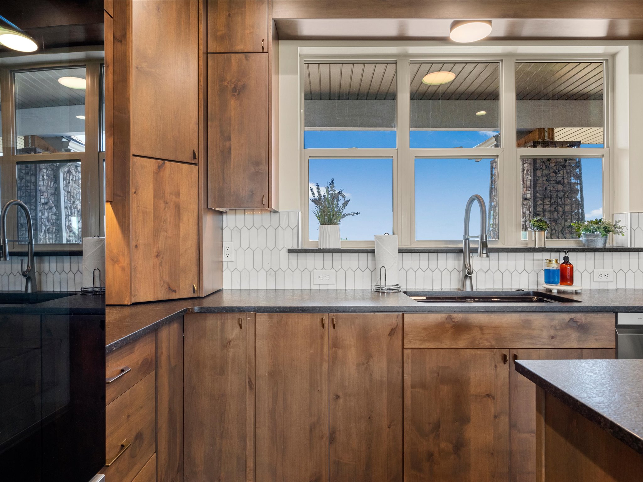 Kitchen featuring wood cabinetry, dark countertops, a window overlooking a natural rock feature, a gooseneck faucet, and a white picket tile backsplash