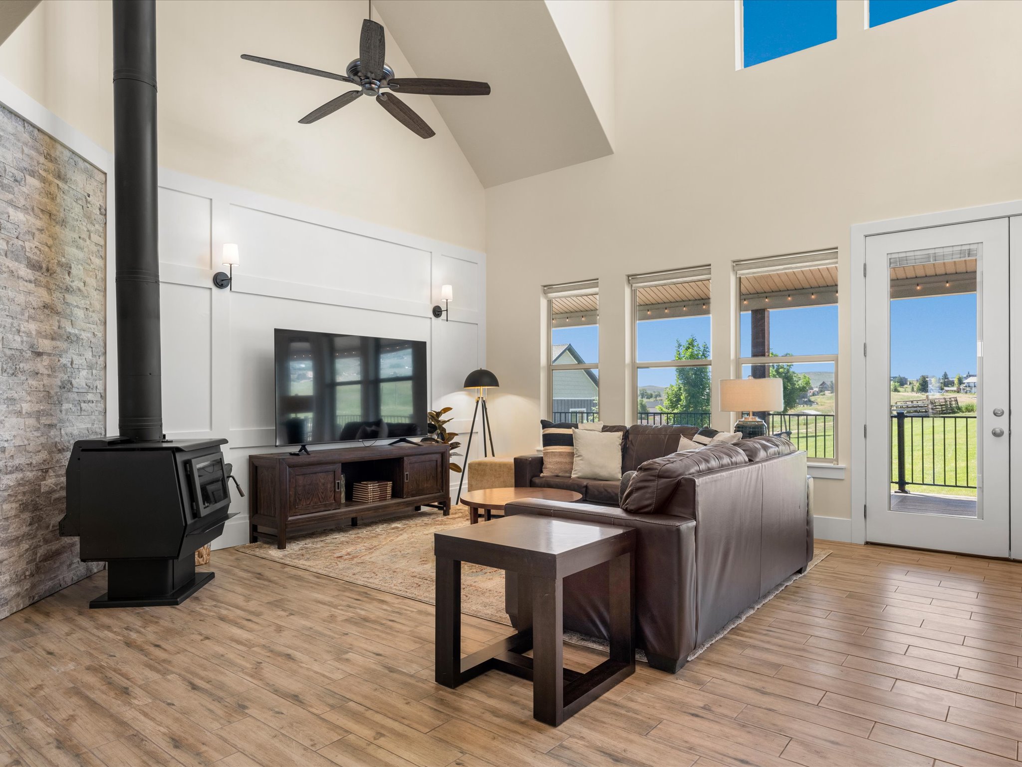 Vaulted living area featuring a black wood-burning stove with a stone-clad wall accent, wood-finish tile flooring, and multiple windows