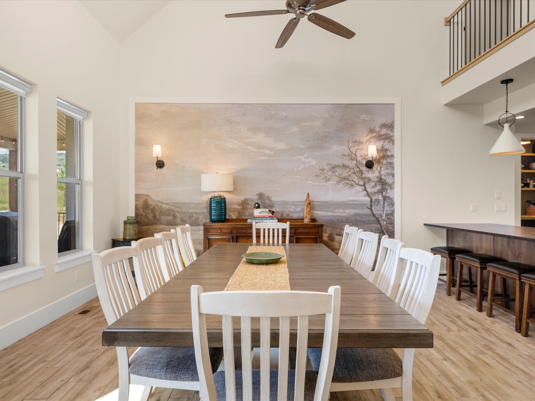 Vaulted ceiling dining area featuring a decorative mural wall, wood-finish tile flooring, two large windows, and a ceiling fan