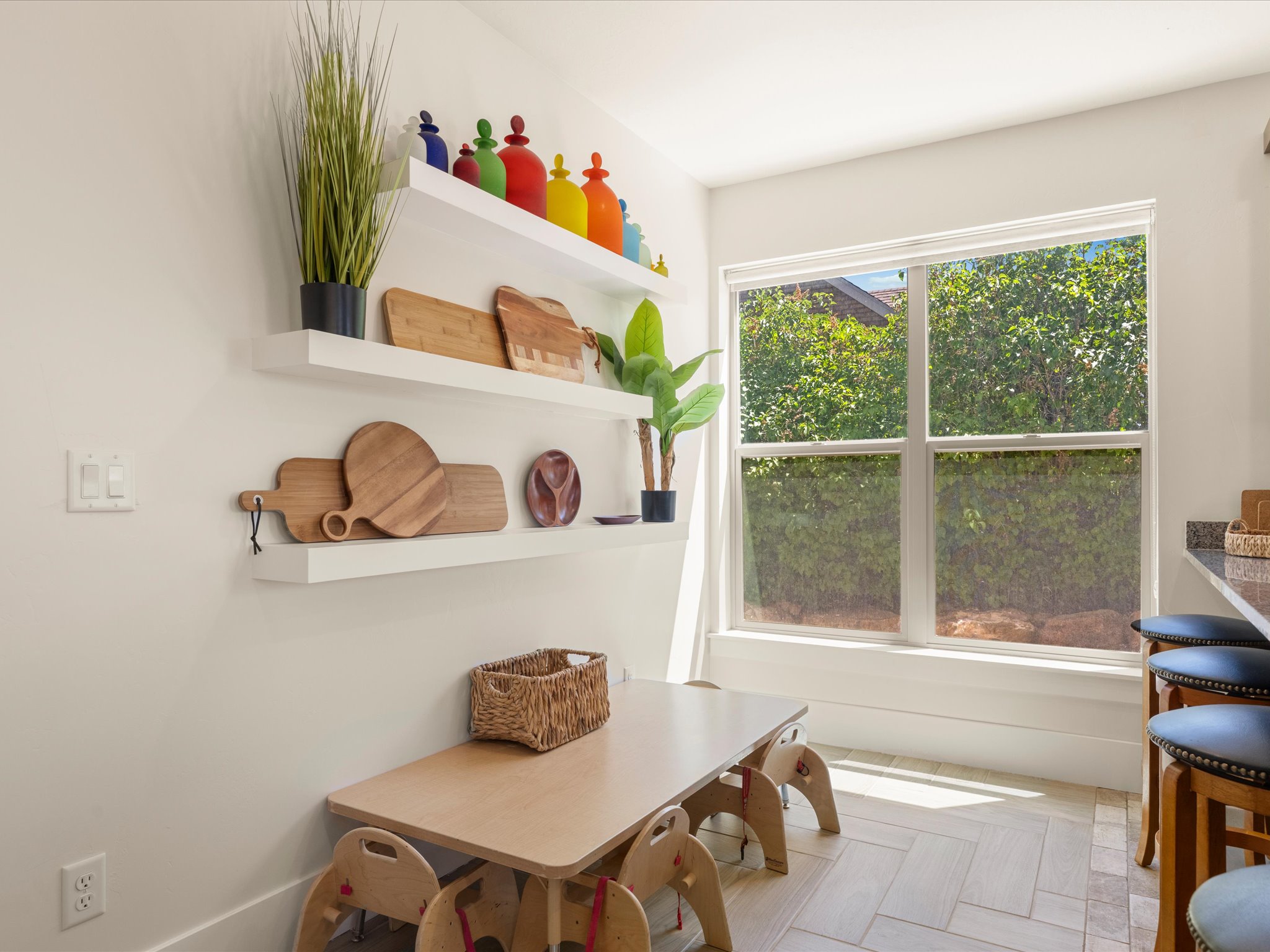 Bright dining area featuring a large window, white floating shelves, and herringbone-patterned tile flooring