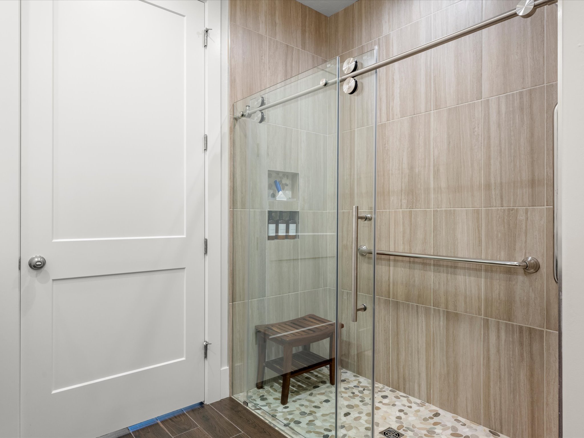 Walk-in shower featuring a frameless glass enclosure, natural stone pebble flooring, wood-finish wall tiling, a built-in shower niche, and a brushed nickel grab bar