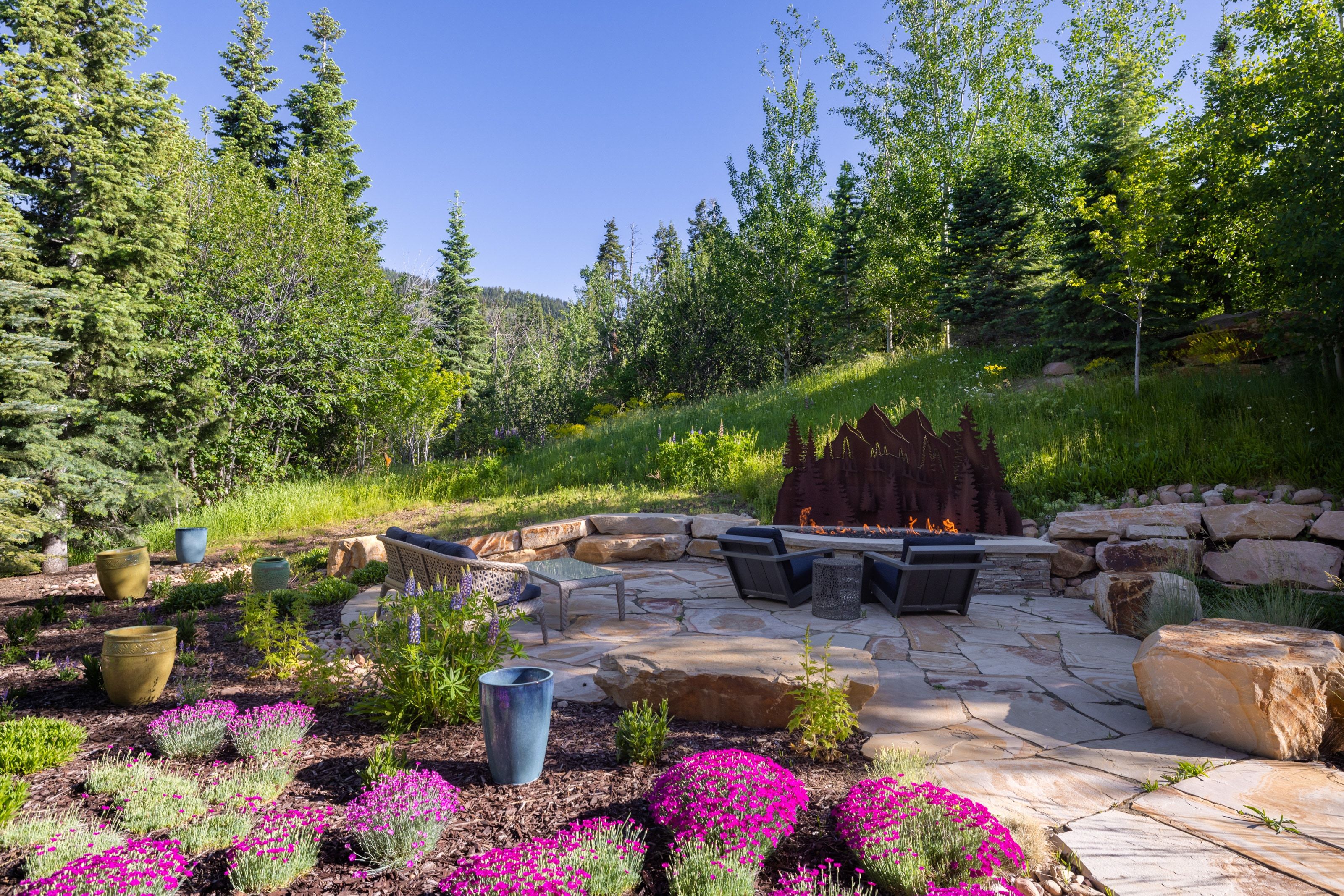 Outdoor living area featuring a flagstone patio, built-in fire pit with decorative metal surround, natural stone retaining walls, and planted garden beds with flowering ground cover