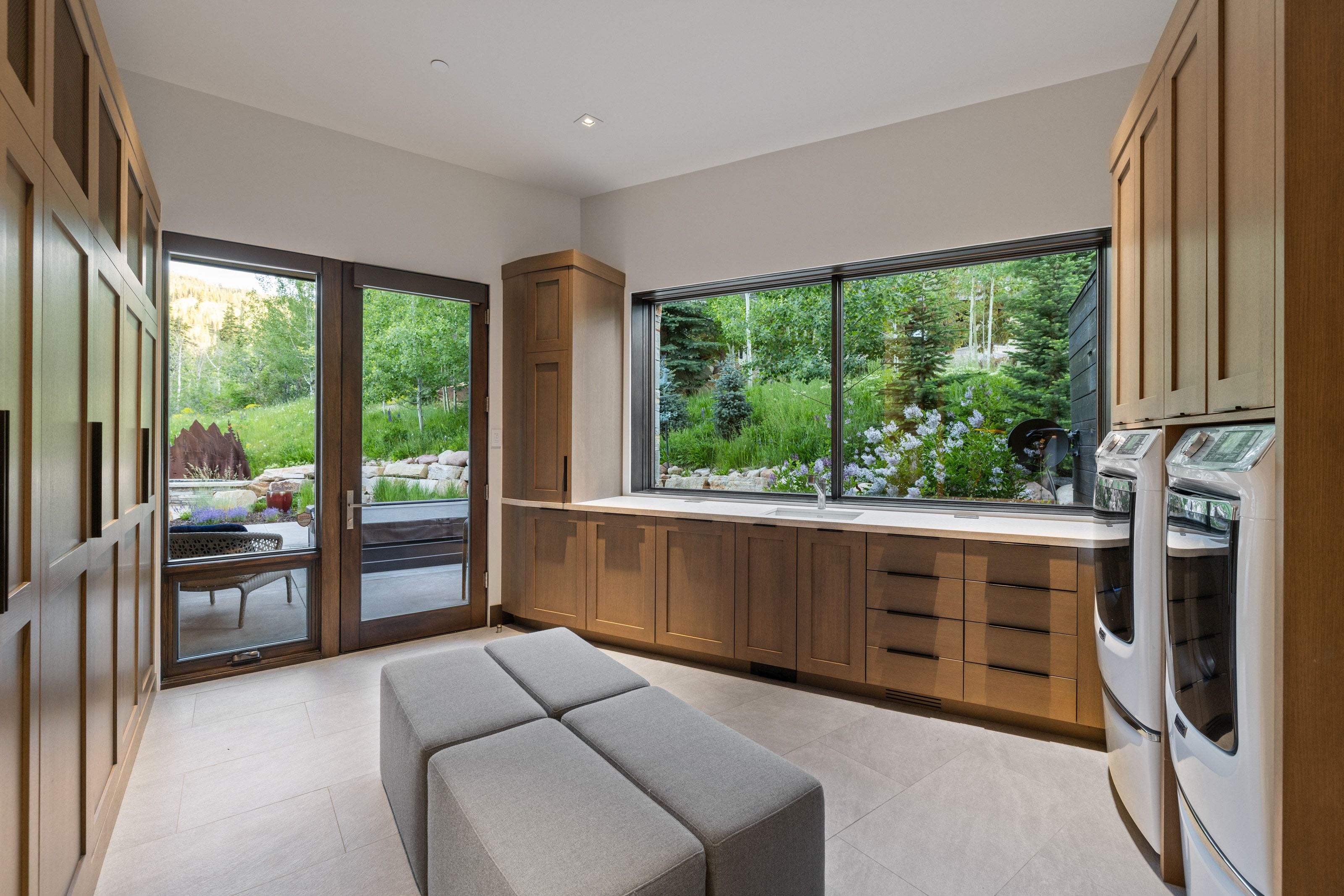 Spacious utility room featuring extensive built-in wooden cabinetry, a wide horizontal window, dual glass-paneled doors, and light-toned tile flooring