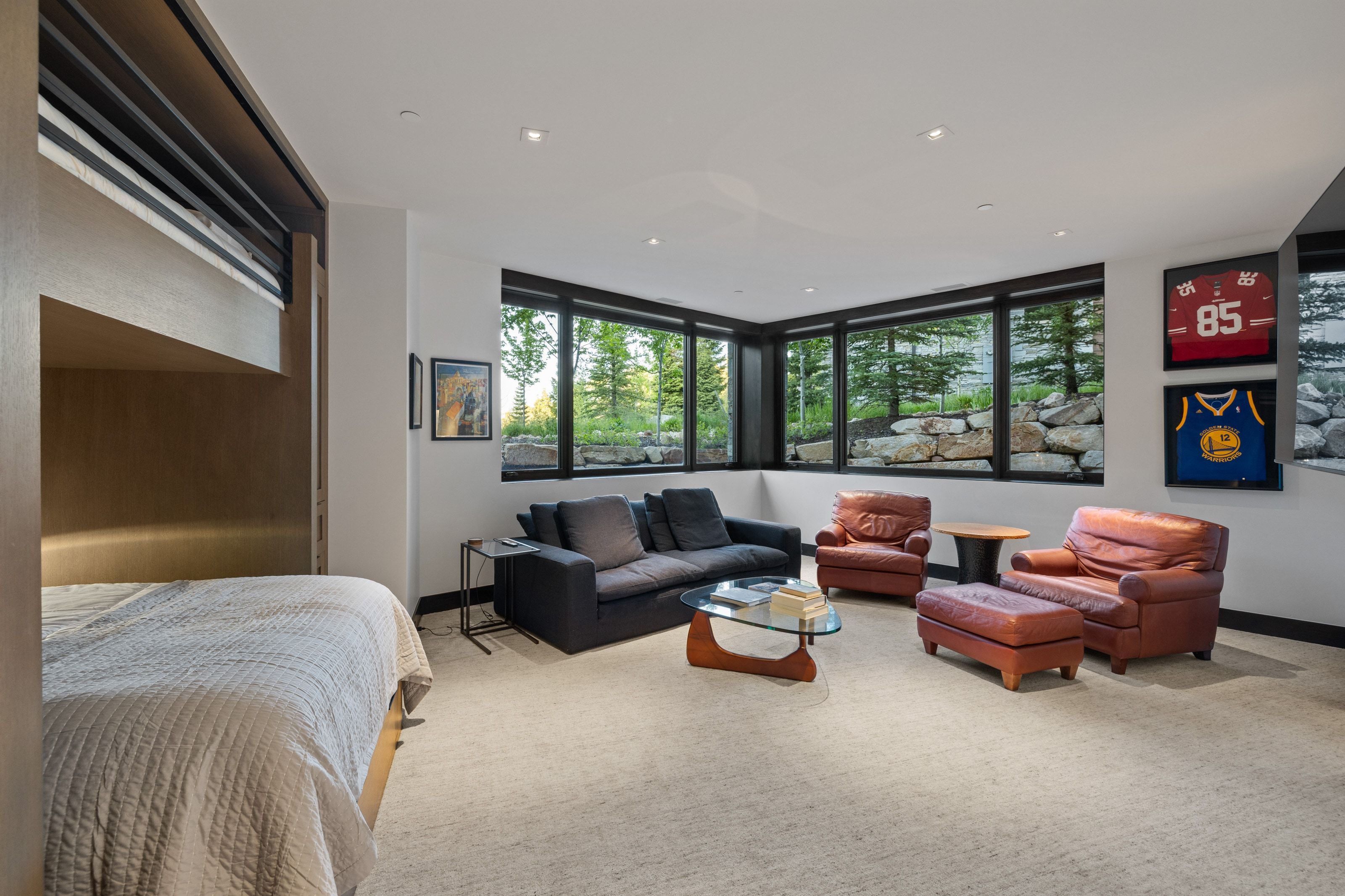 Recessed lighting illuminates a spacious room featuring a built-in wood-finish bunk bed, wall-to-wall carpeting, and expansive windows showcasing exterior landscaping