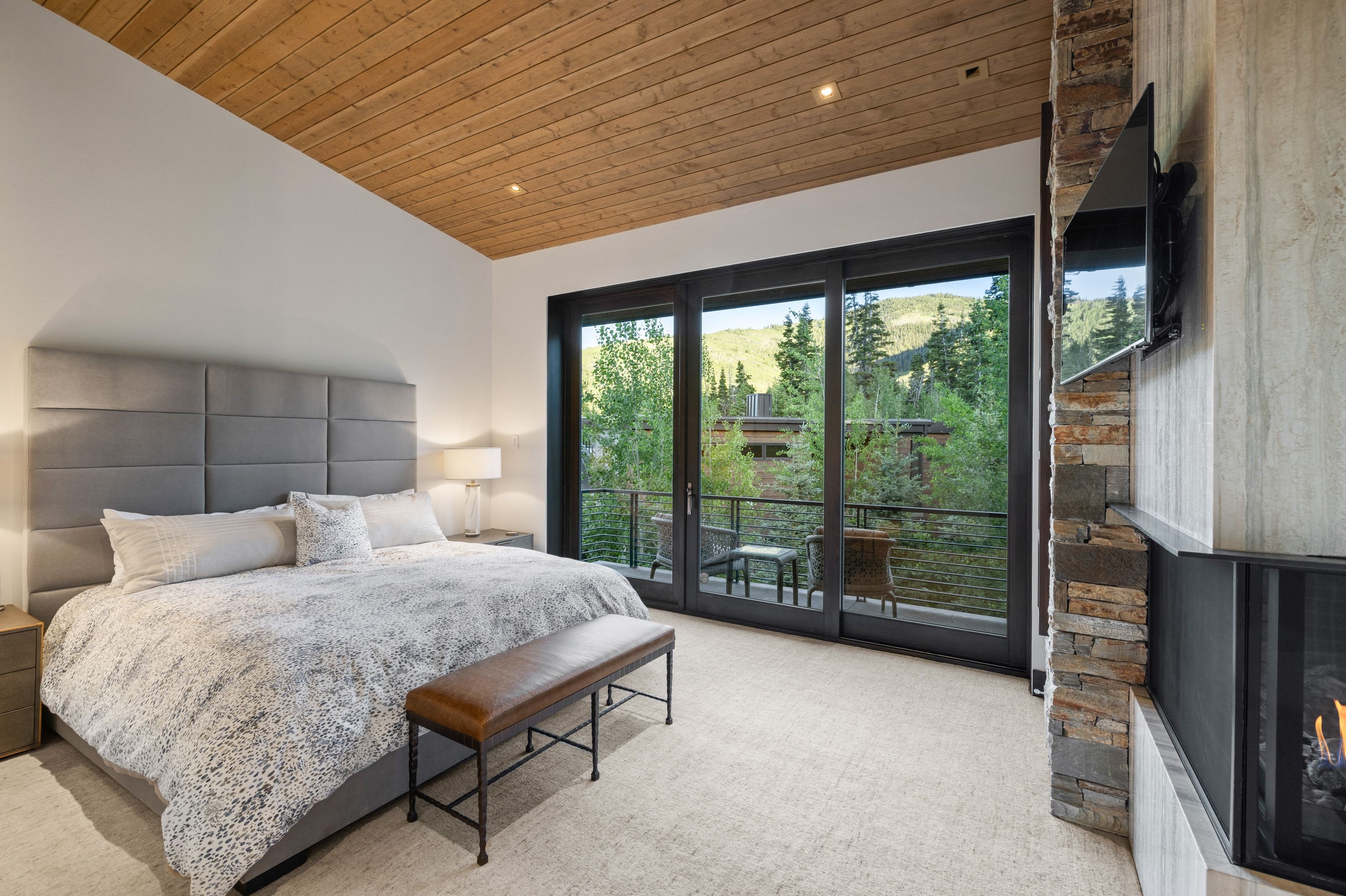 Recessed lighting illuminating a wood-paneled ceiling, sliding glass doors opening to a balcony, a stone-clad fireplace with a built-in firebox, and neutral carpeting