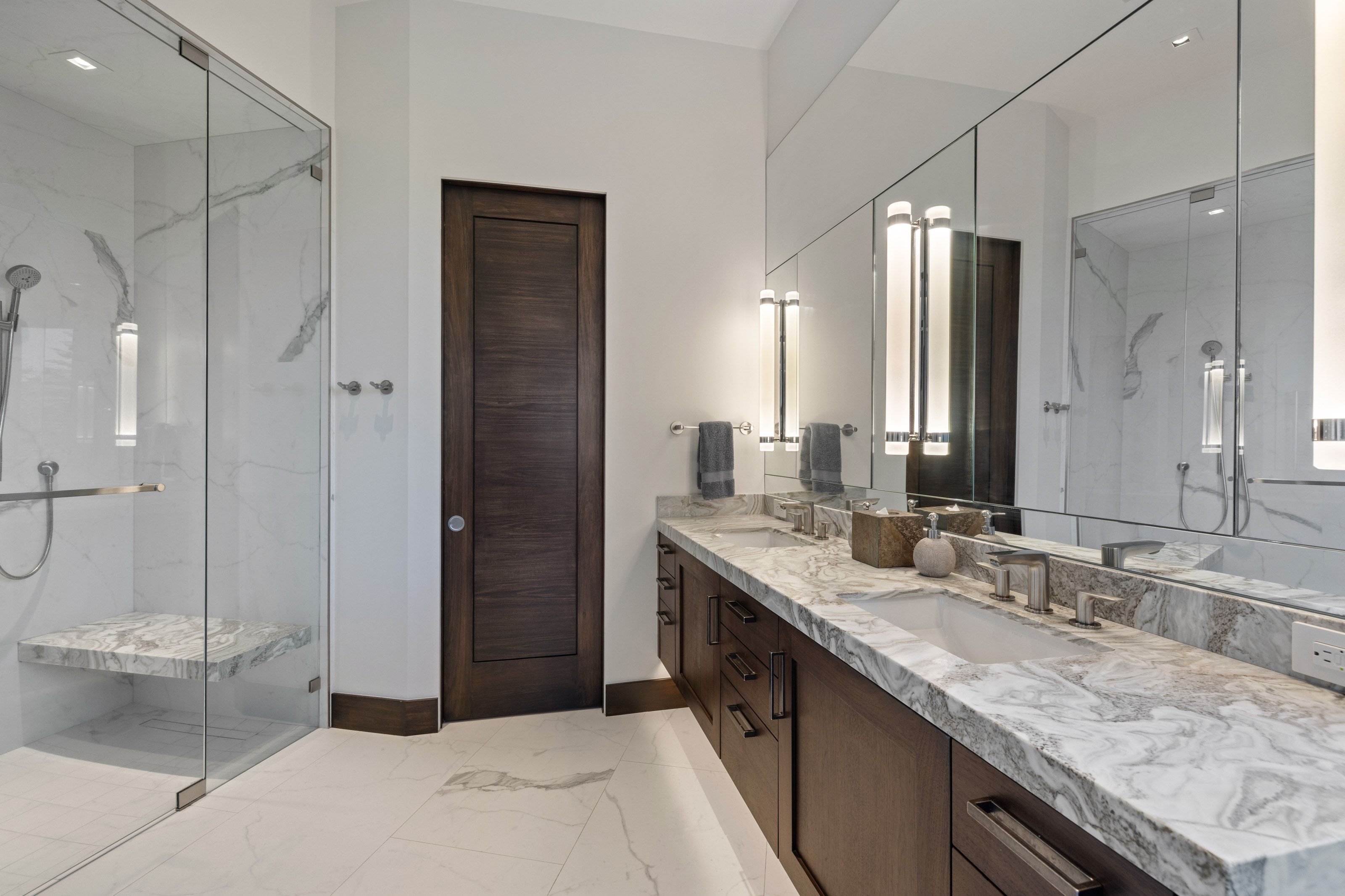 Spacious bathroom featuring a glass-enclosed shower with a built-in bench, dual vanity with undermount sinks, stone countertops, and integrated wall mirrors