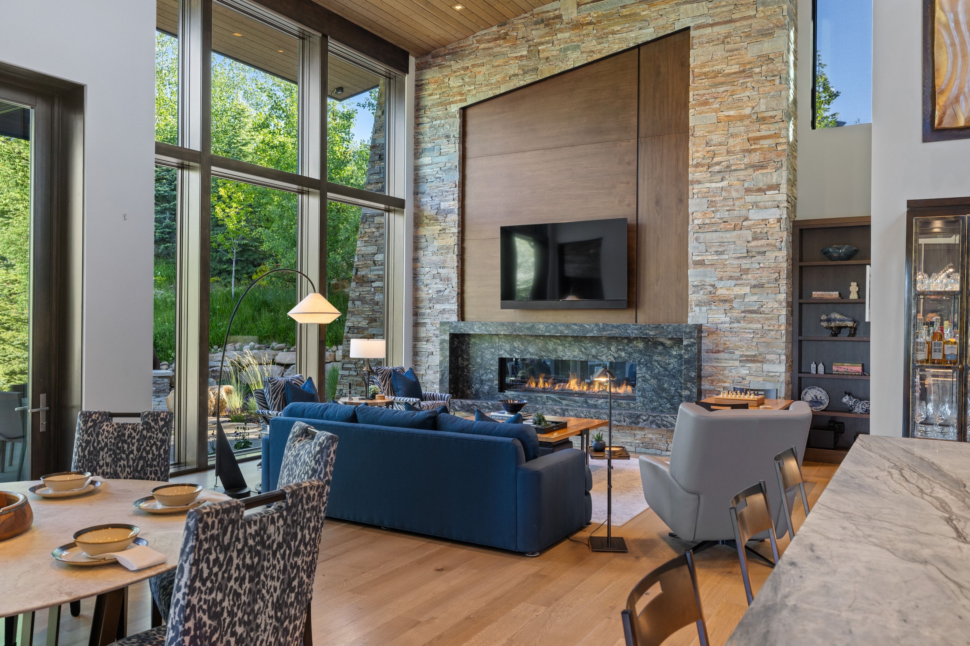 Two-story window wall, stacked stone fireplace, linear hearth, wood-finish flooring, and natural wood ceiling