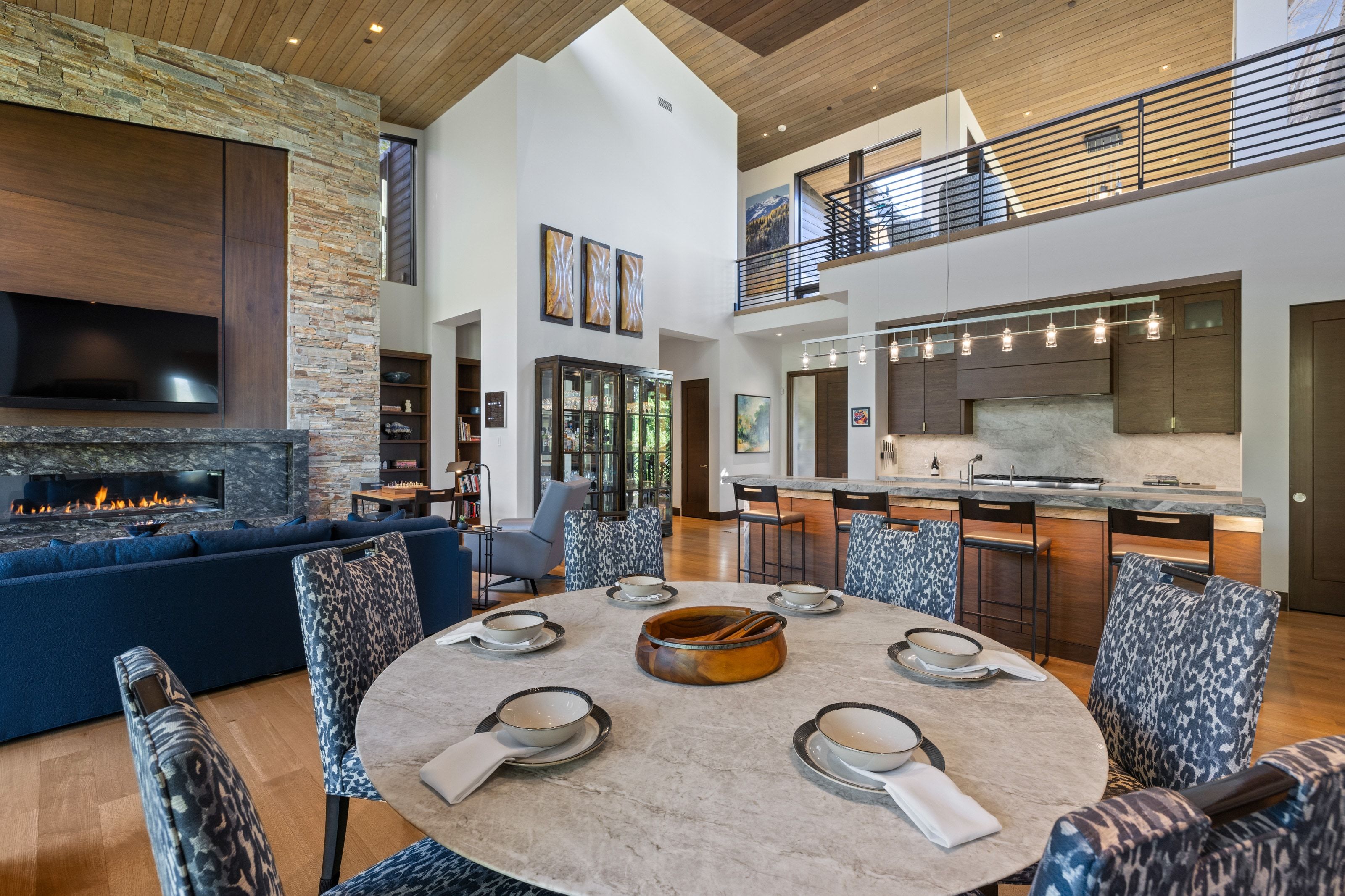 Open-concept living space featuring a high vaulted wood-paneled ceiling, a stone fireplace with a linear firebox, a second-story interior balcony with metal railing, an island kitchen with dark wood-finish cabinetry, and wood-finish flooring