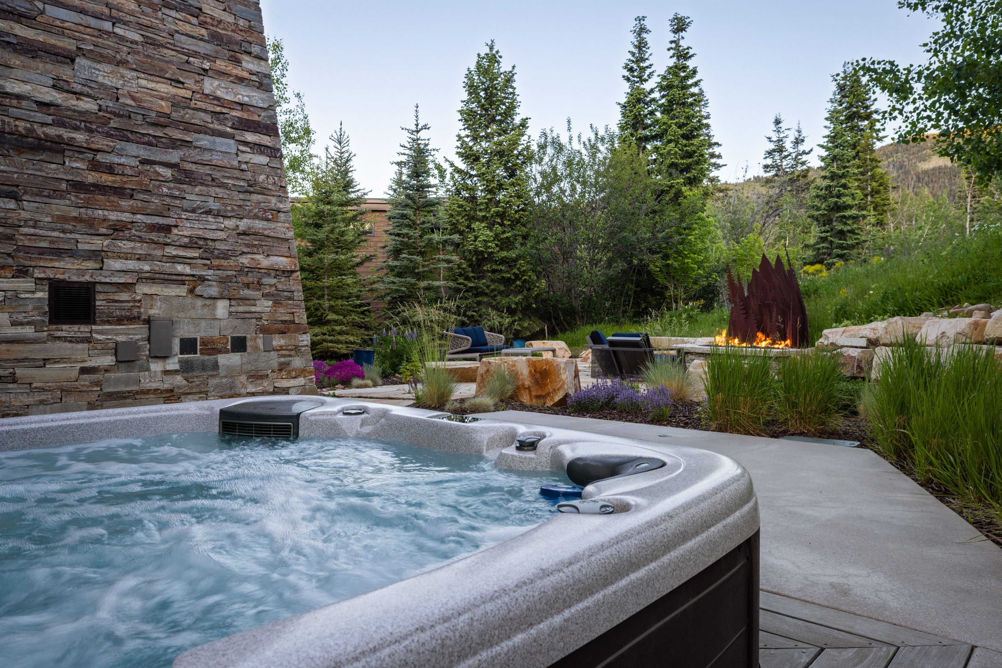 Bubbling spa bordered by a stacked stone wall, a concrete pathway, and mature evergreen trees