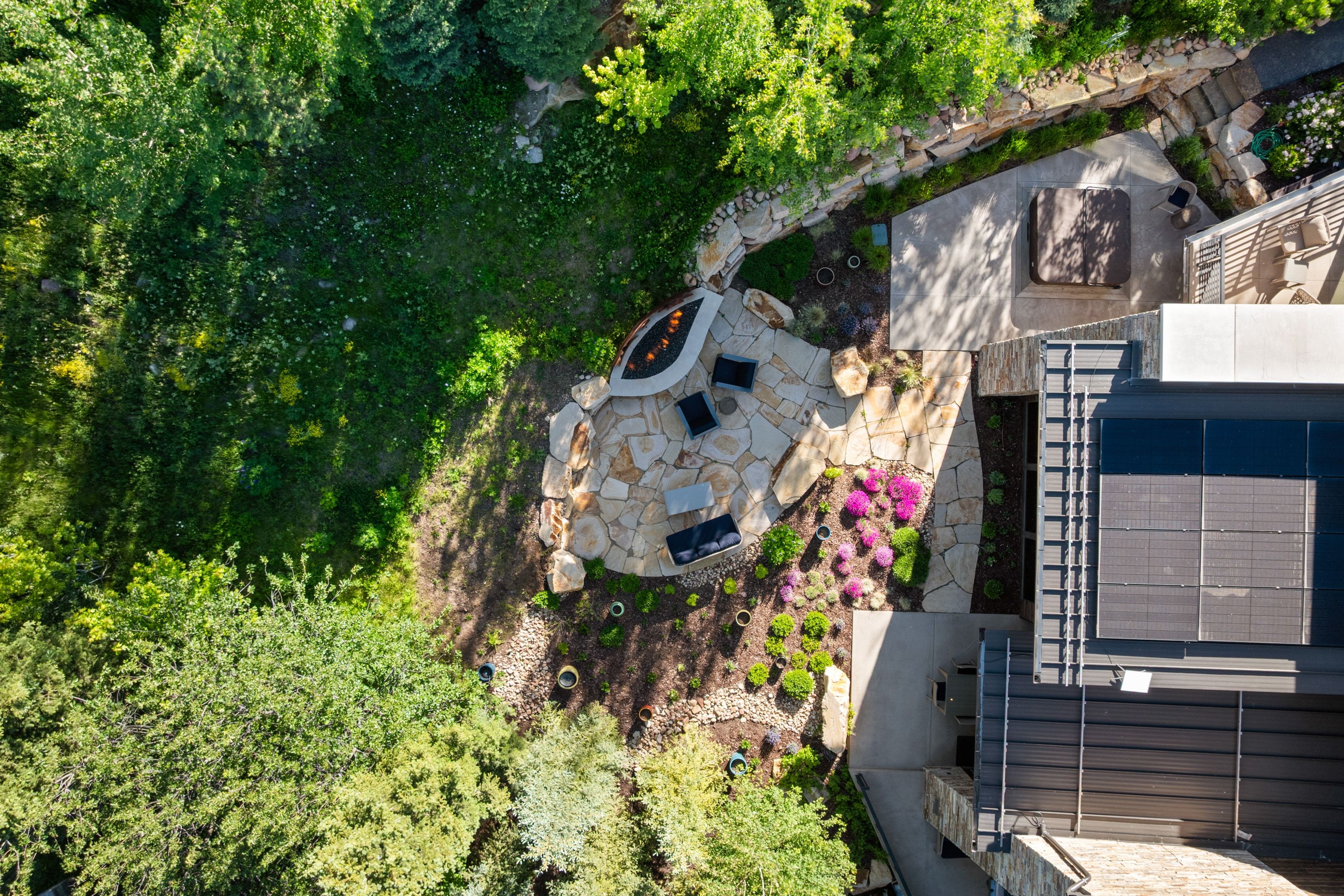 Expansive stone patio featuring a built-in fire pit, surrounded by mature trees and vibrant landscaping