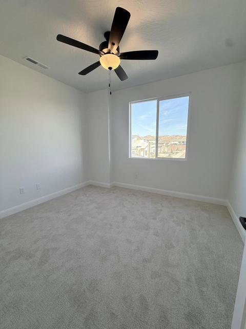 Bright room featuring a large window, plush gray carpet, clean white walls, a ceiling fan with integrated lighting, and white baseboards