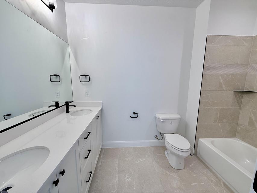 Contemporary bath featuring a dual vanity with white cabinetry and integrated sinks, matte black fixtures, an expansive wall mirror, and a bathtub with tiled surround