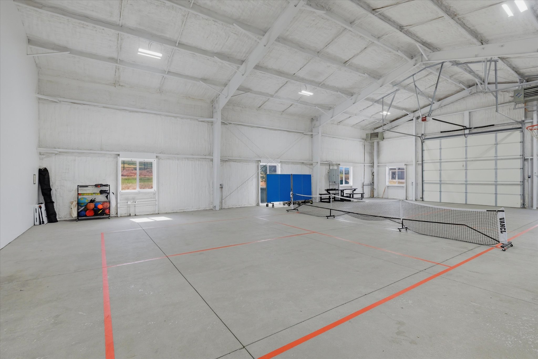 Spacious indoor court featuring high ceilings with exposed trusses, concrete flooring with painted lines, multiple windows, and a large roll-up door